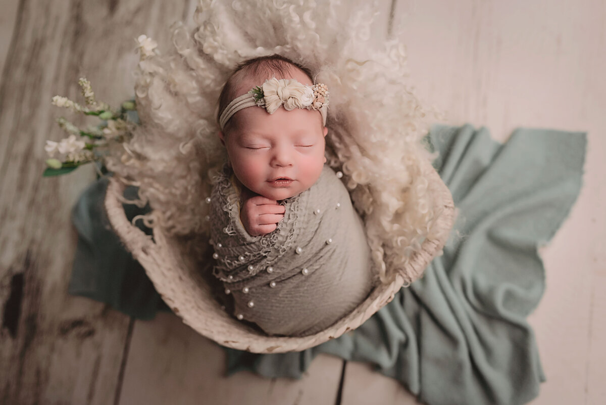 newborn baby girl in sage green wrap with pearls in white basket on white wood flooring with beige and florals