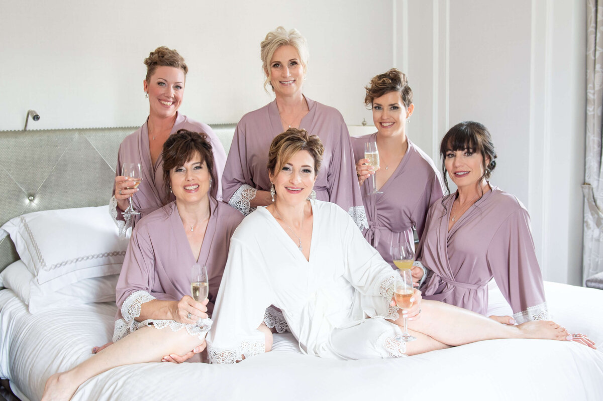 an elegant Chateau Laurier wedding bride sits on the bed in the Prime Minister's Suite with her bridesmaids around her in pretty pink robes.  Captured by Ottawa wedding photographer JEMMAN Photography