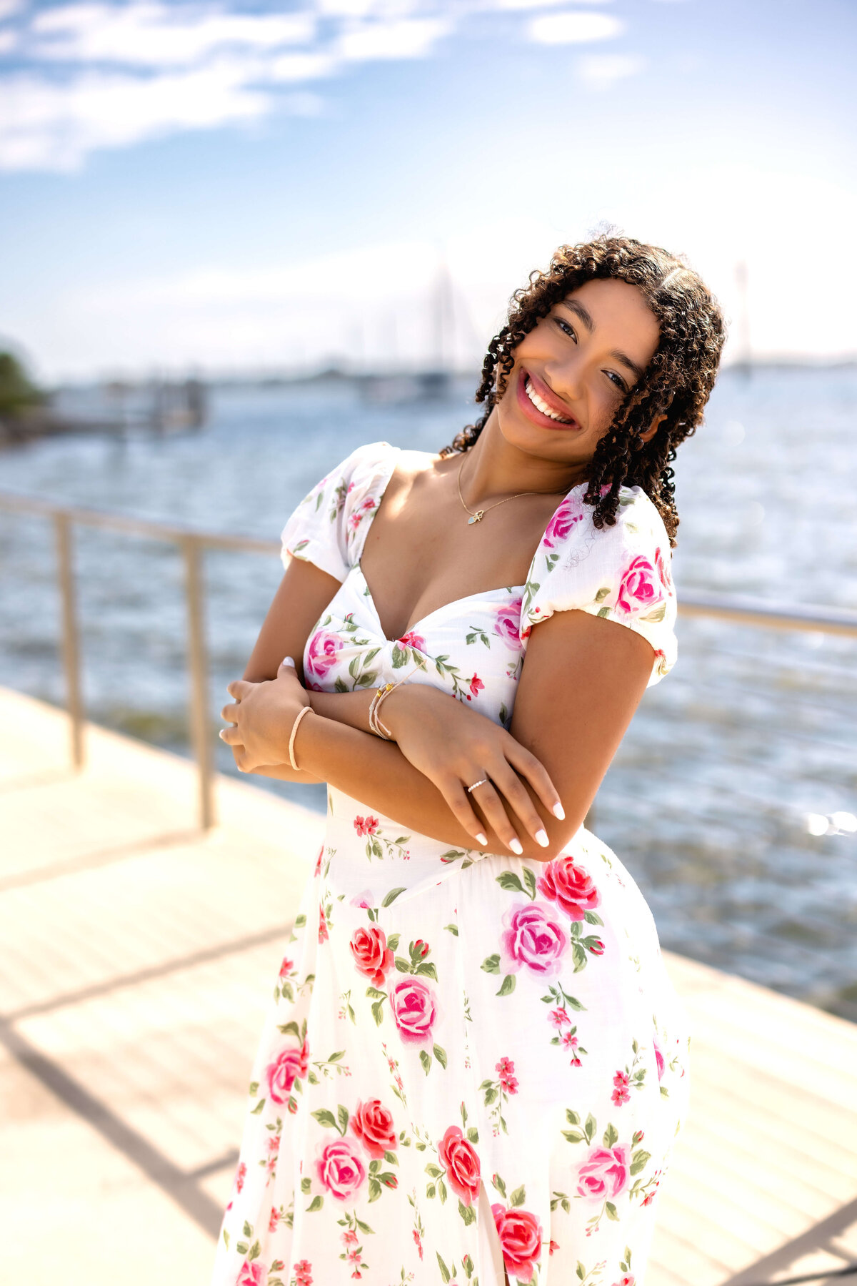 Close-up Lake Nona senior portrait of a girl with arms crossed, smiling brightly in a floral dress, soft ocean backdrop behind her.