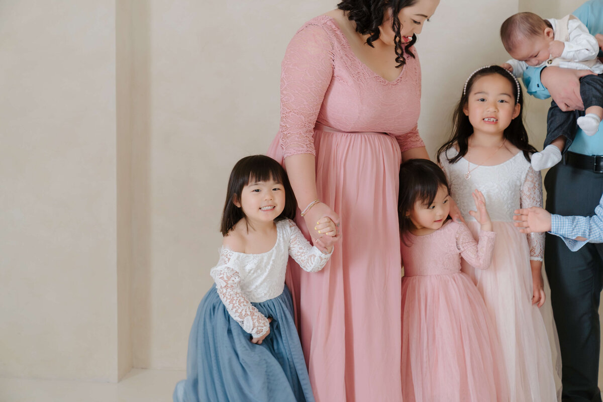Mother holding hands with her daughters while standing together in soft pink and cream tones at the studio.