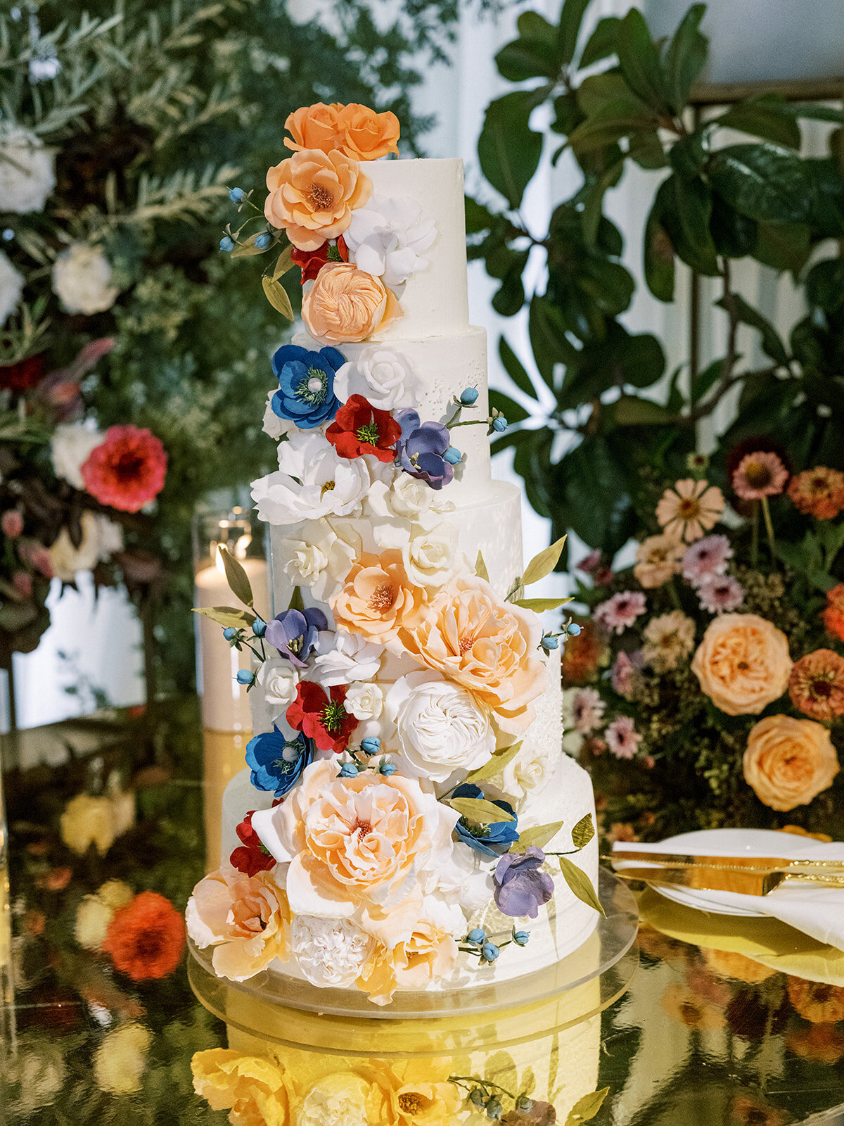 Elegant four-tier wedding cake adorned with peach, blue, and red sugar florals—artful, vibrant, and full of celebration charm.