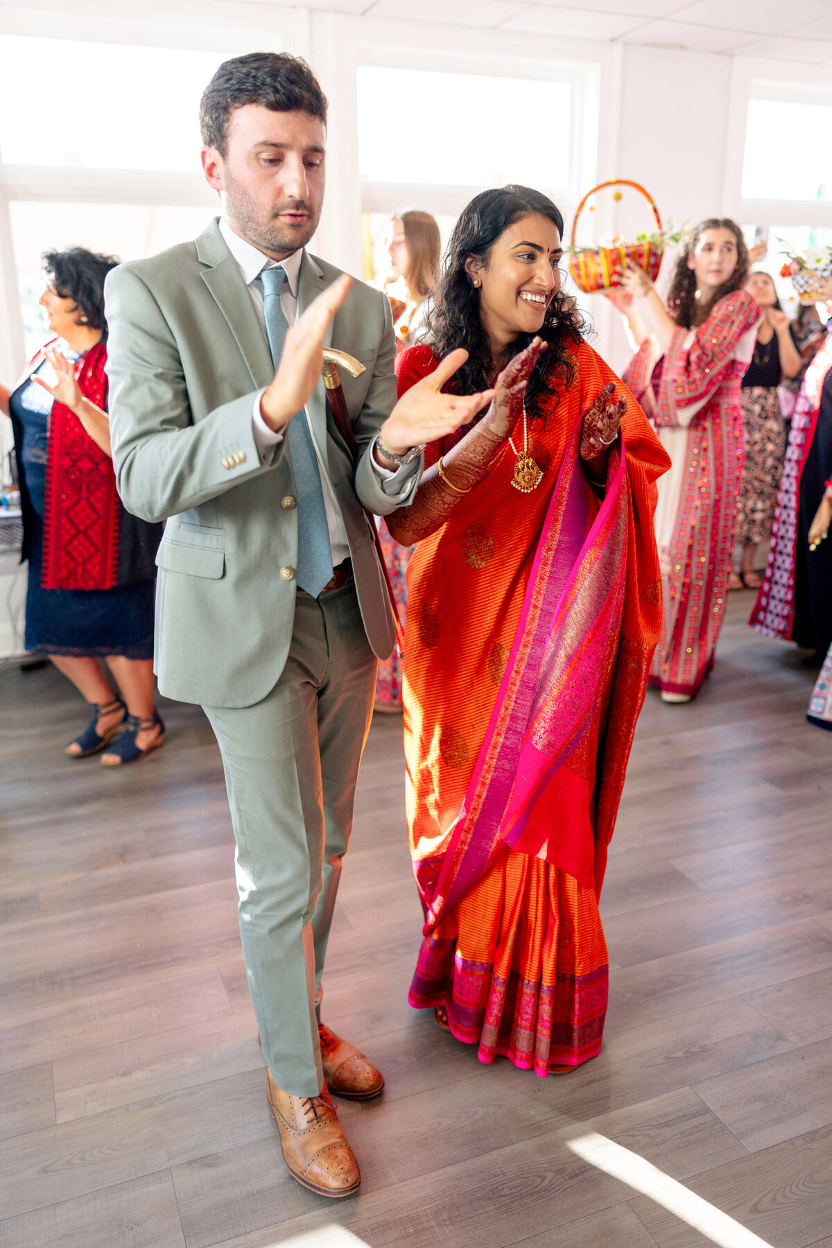 A man in a gray suit and a woman in a vibrant red sari are clapping and dancing at a festive indoor gathering. Other people in colorful attire can be seen in the background, creating a lively and joyful atmosphere.
