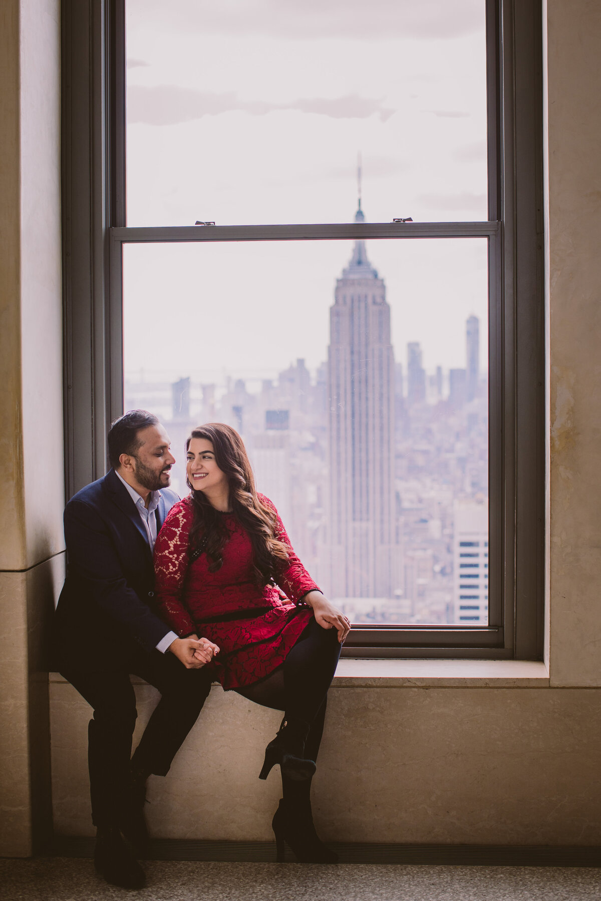 A couple sitting by the window sill at the top of the rock with the view of the empire state building