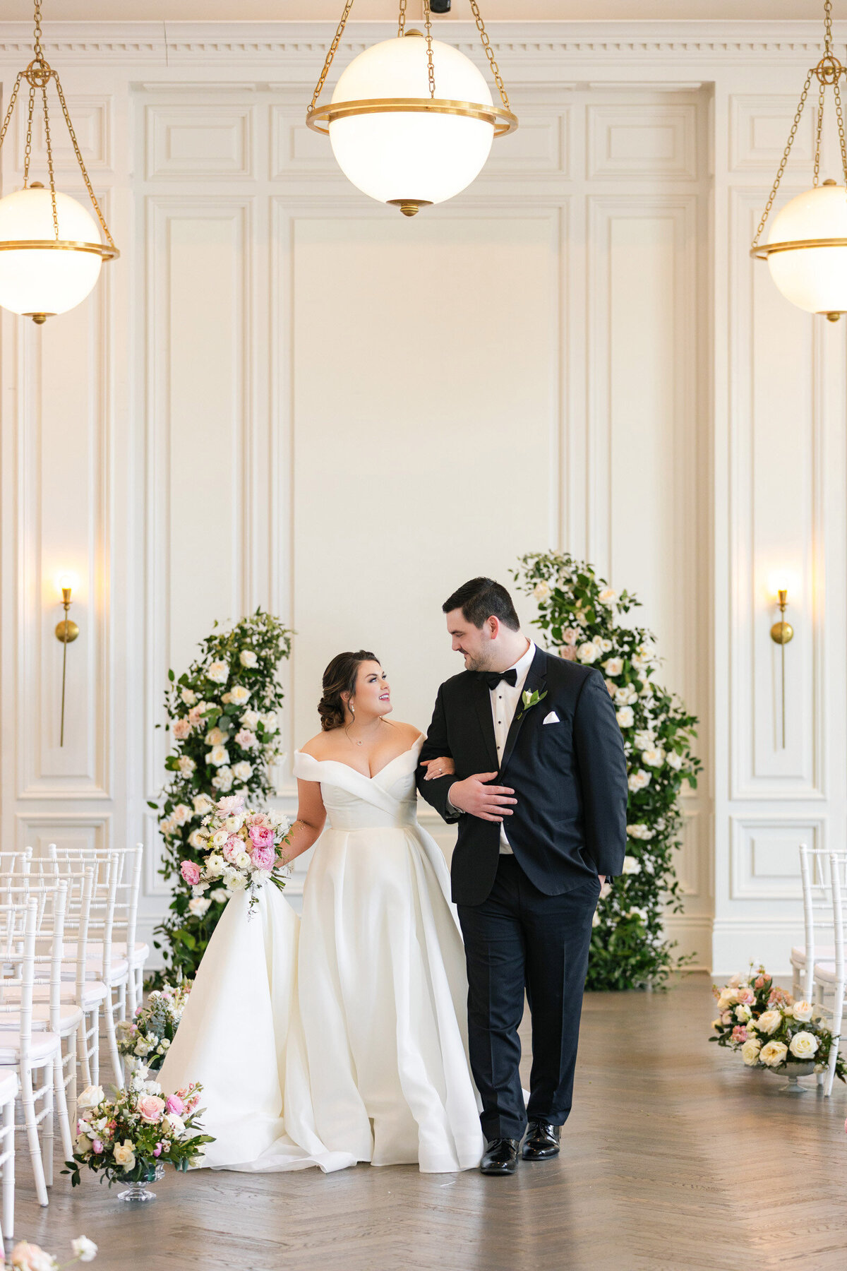 horizontal portrait of the couple walking down the aisle in the Governor’s Room at The Adolphus in Dallas, with a floral arch behind them and looking at each other, capturing a romantic and elegant wedding moment.