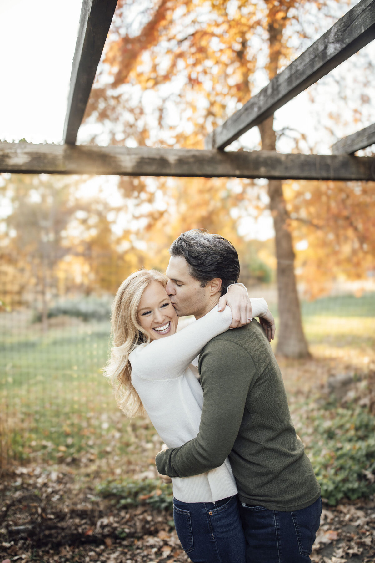 Couple sharing cheek kiss among colorful trees during session at Cross Estate Gardens in Bernardsville Somerset County New Jersey