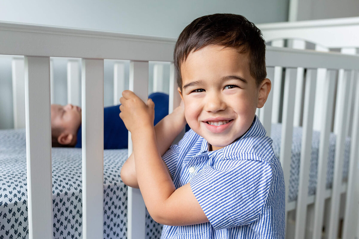A boy standing in front of his newborn brother's crib smiling during a newborn session in Ashburn, VA.