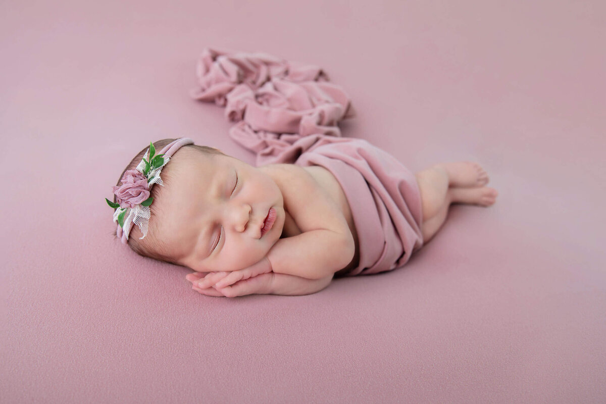 Newborn baby girl laying on her side wrapped in pink, headband and background for their Burlington newborn photography session.