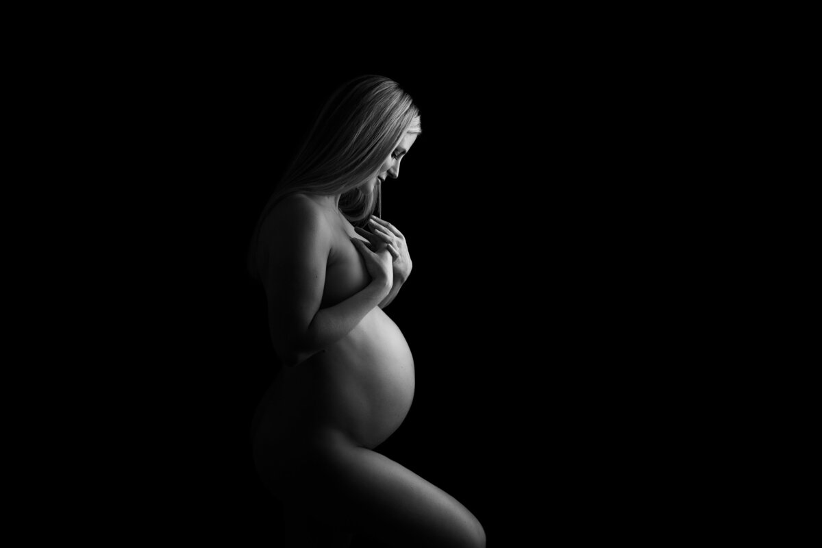 Nude maternity portrait on a black background with a woman lovingly looking down at her belly with her hands placed over her breasts.