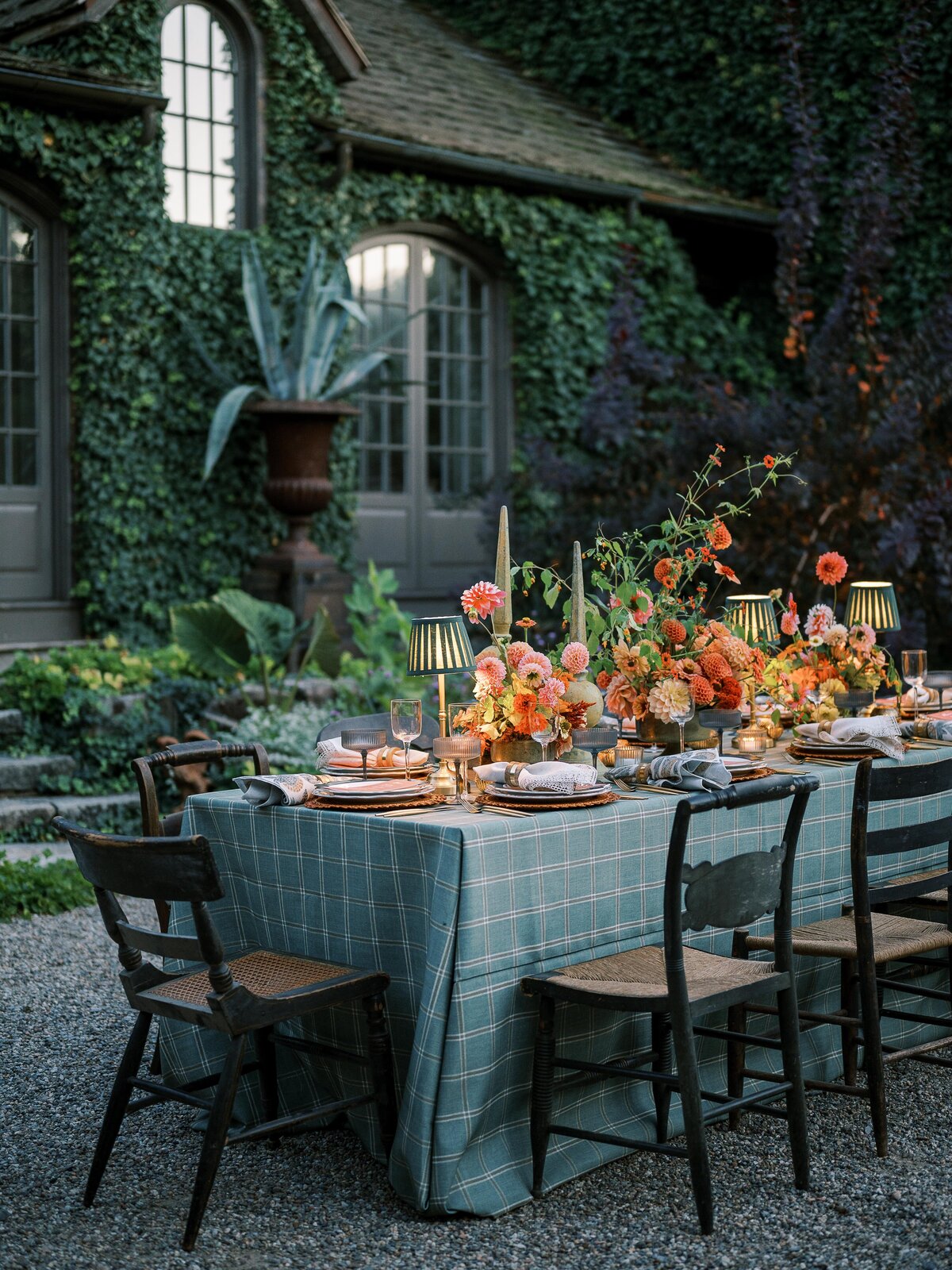 An outdoor reception table with a blue plaid tablecloth, adorned with lush floral arrangements in warm tones, set in a courtyard with ivy-covered walls.