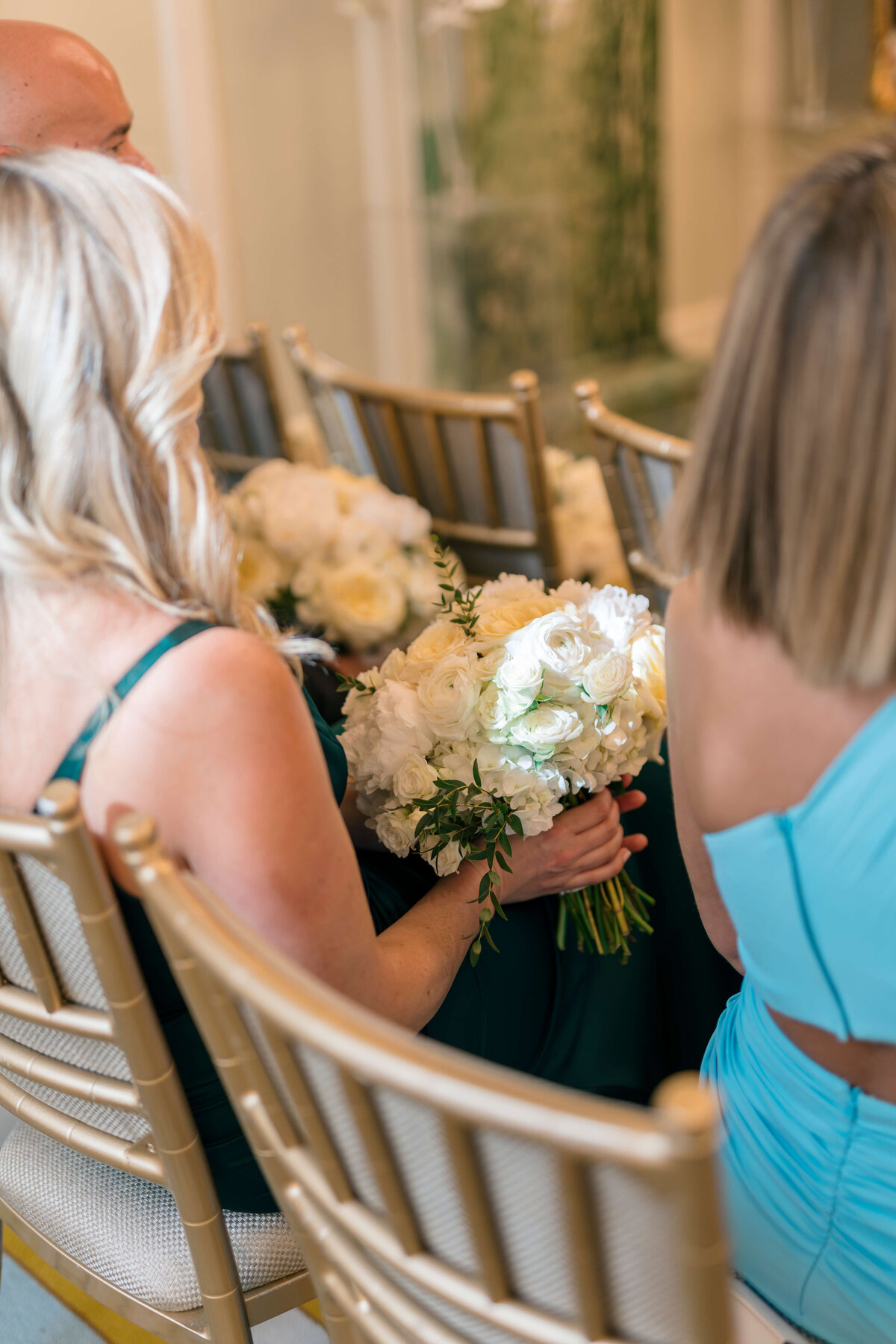 A woman with blonde hair sits on a gold chair holding a bouquet of white flowers. She is wearing a dark green dress. Another woman in a light blue dress is seated next to her. The setting appears to be a formal event or ceremony.