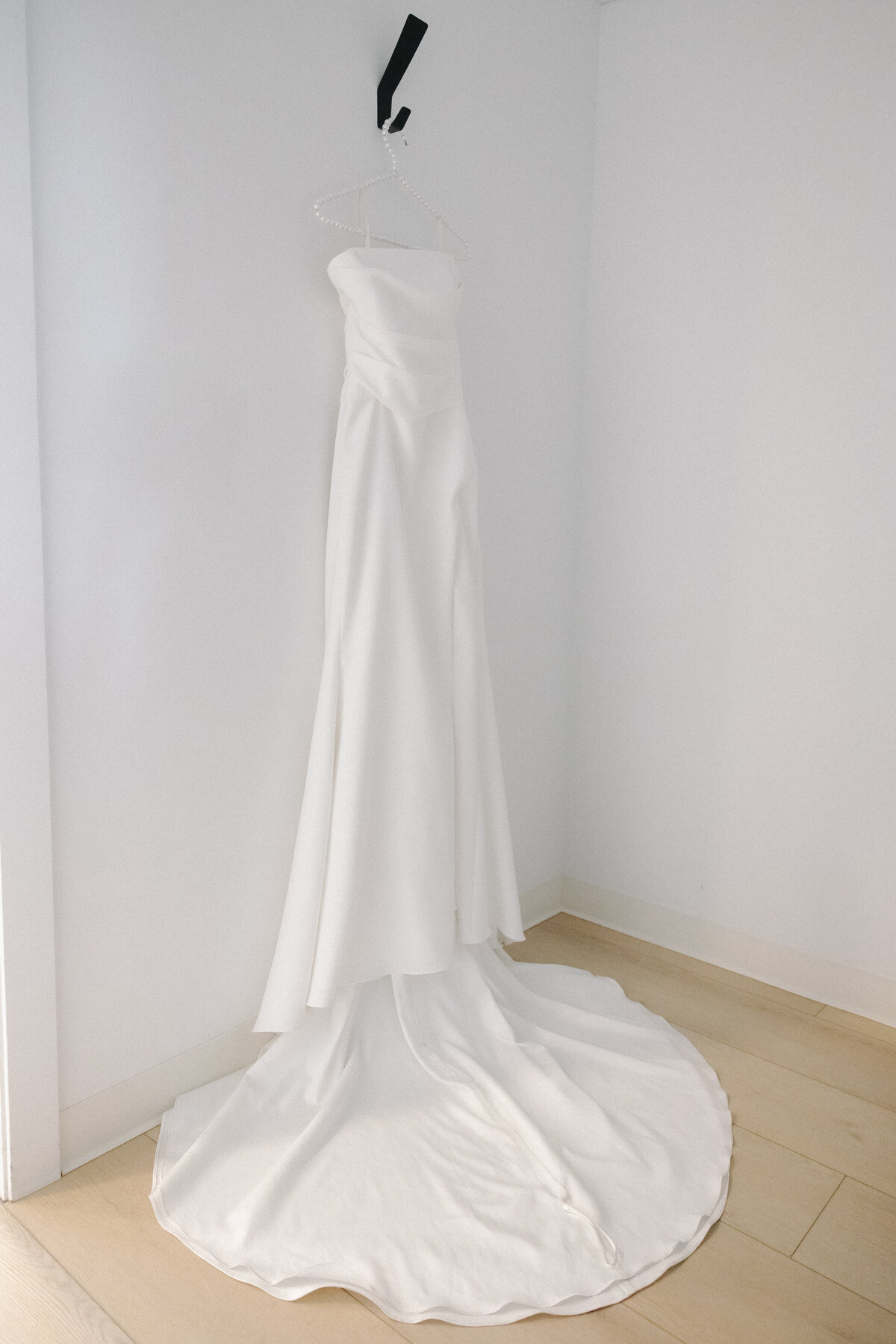 A white satin wedding dress hanging from a thick black hook. The hanger holding the dress has white pearls on it. The dress is flowey with pleats toward the top and the train is long laid out nicely in a semi circle below the dress on the floor. Natural light comes in through a doorframe to the left of the dress. The walls are white and the floor is a washed out wooden laminate. 
