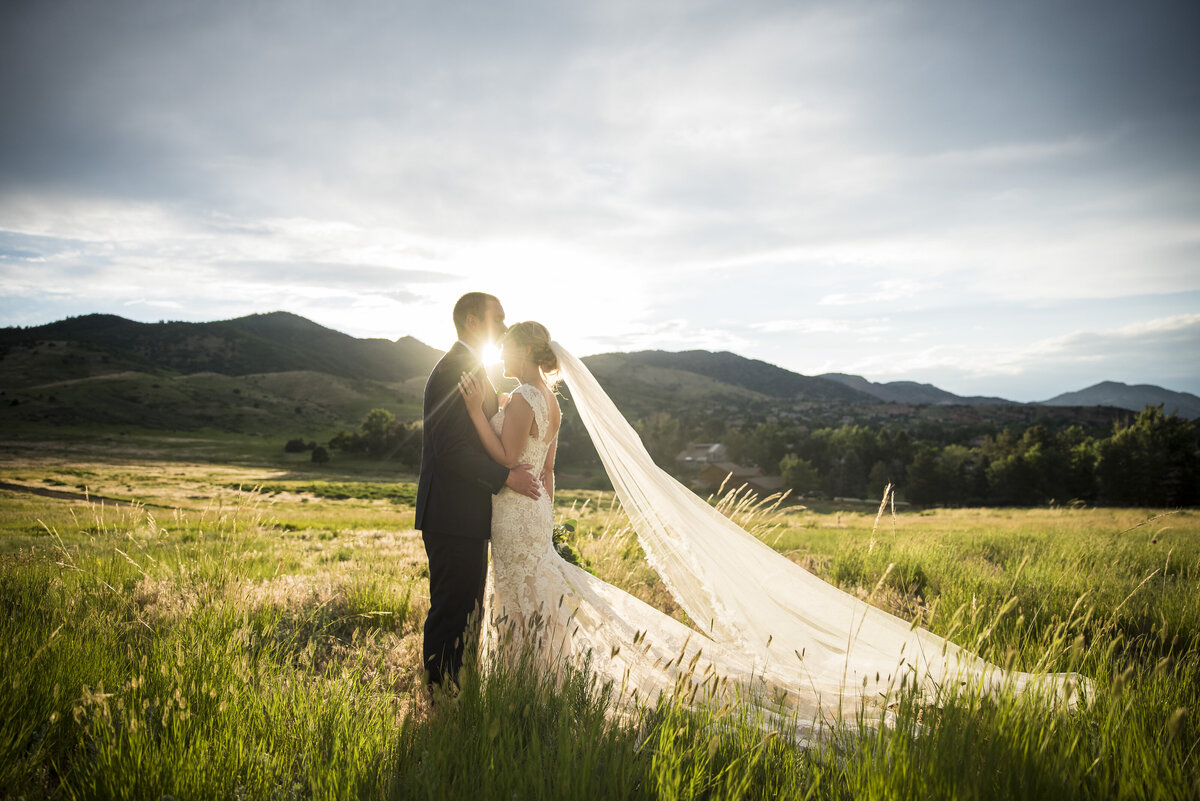 A groom kisses his bride's forehead as the two stand in a field at golden hour.
