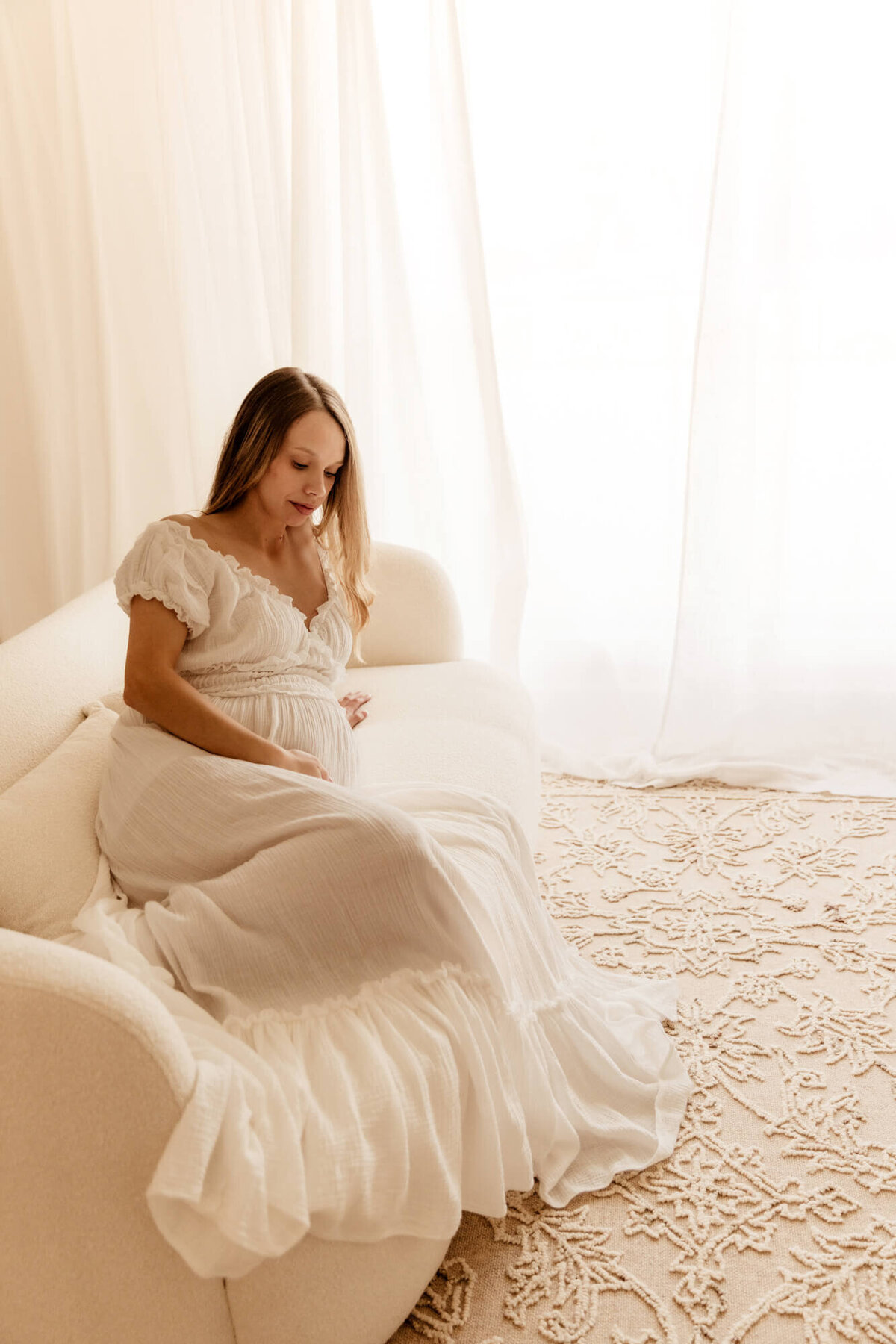 Woman holds her baby bump while sitting on a white couch near a window, Edmond maternity photos.