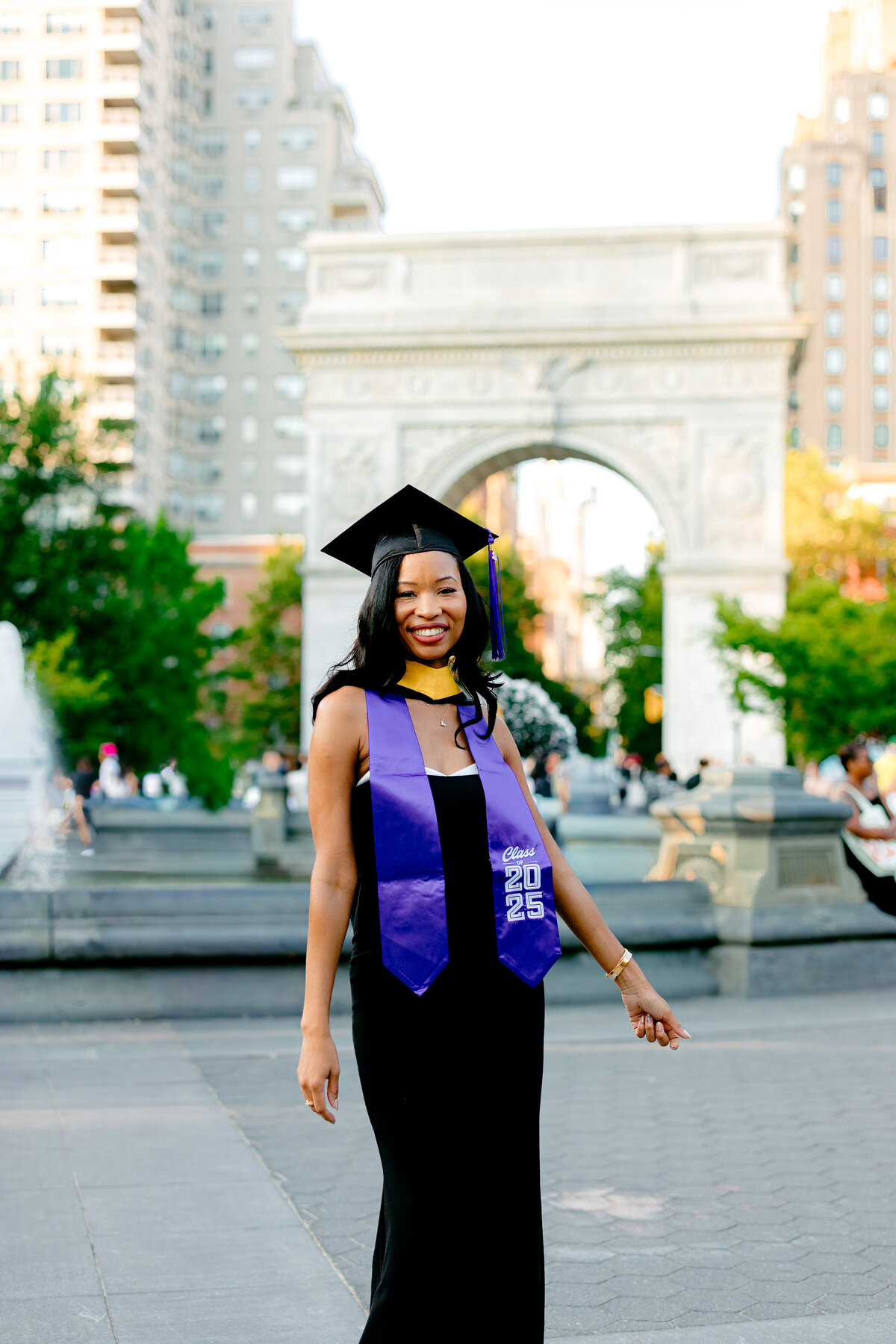 NYU graduate smiling in cap and stole at Washington Square Park during sunset.