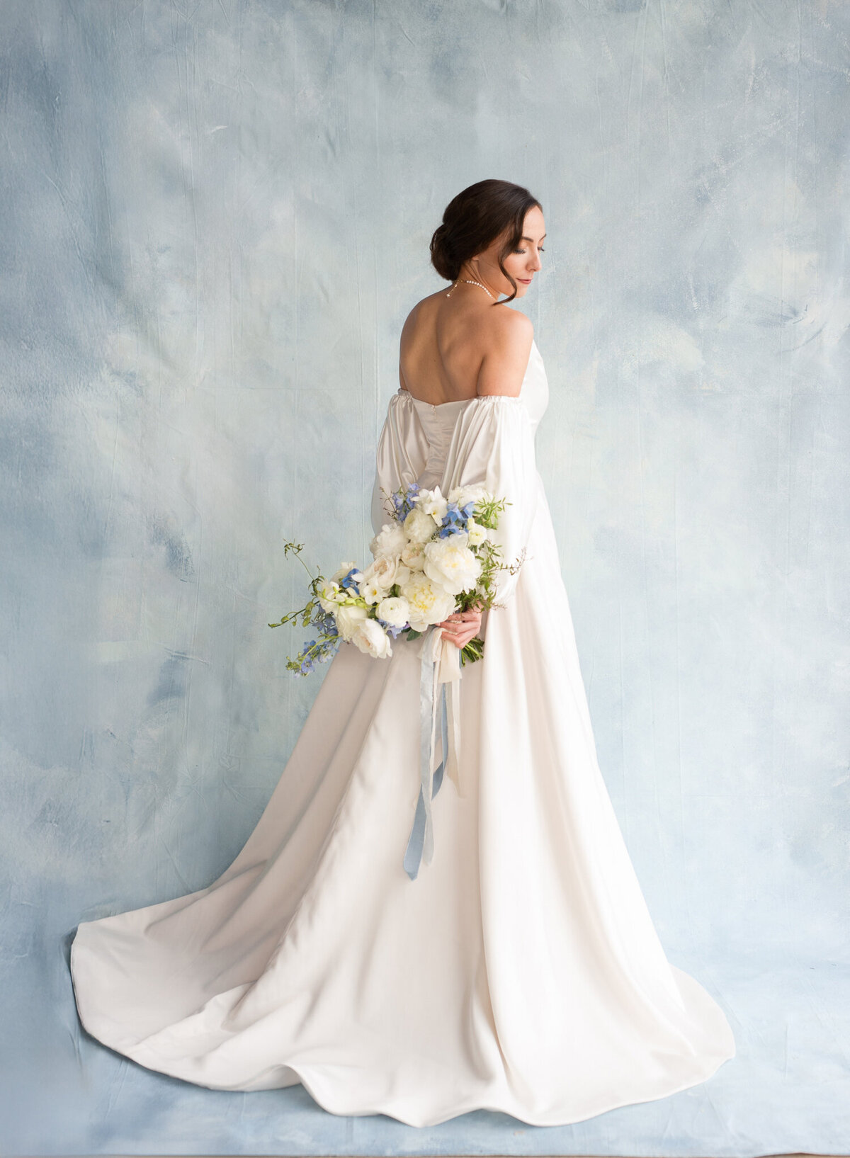 luxury bride holding dusty blue bridal bouquet wearing stephani mai bridal gown in front of blue backdrop