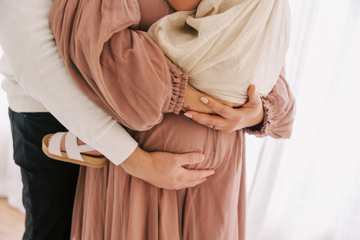 Woman whose husband is standing behind her. She is holding her toddler on her belly and her husband's hand is also on her pregnant belly. They are standing near a window in a portland oregon studio.