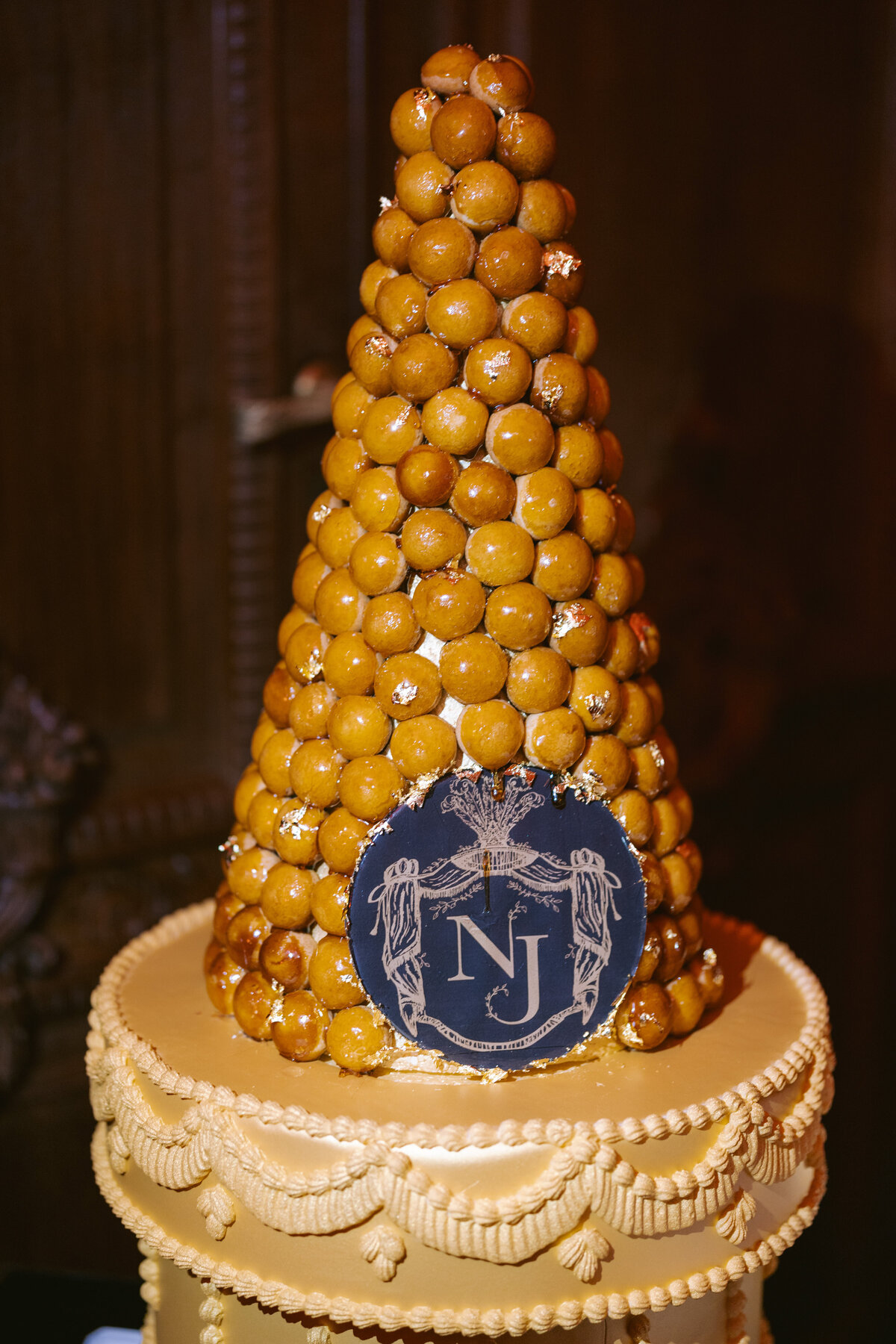 The photo was taken at Shangri La Hotel & Ritz Paris Hotel in Paris, France by photographer Matteo Coltro. In addition, Zen Film Works are the videographers who captured the event. In the image, there is a tall, pyramid-shaped wedding croquembouche made of small, round pastry puffs and adorned with gold accents. The bottom tier features elaborate decorative icing and a round blue plaque with an emblem showcasing the initials NJ. The background is dimly lit, adding a touch of elegance to the overall composition.