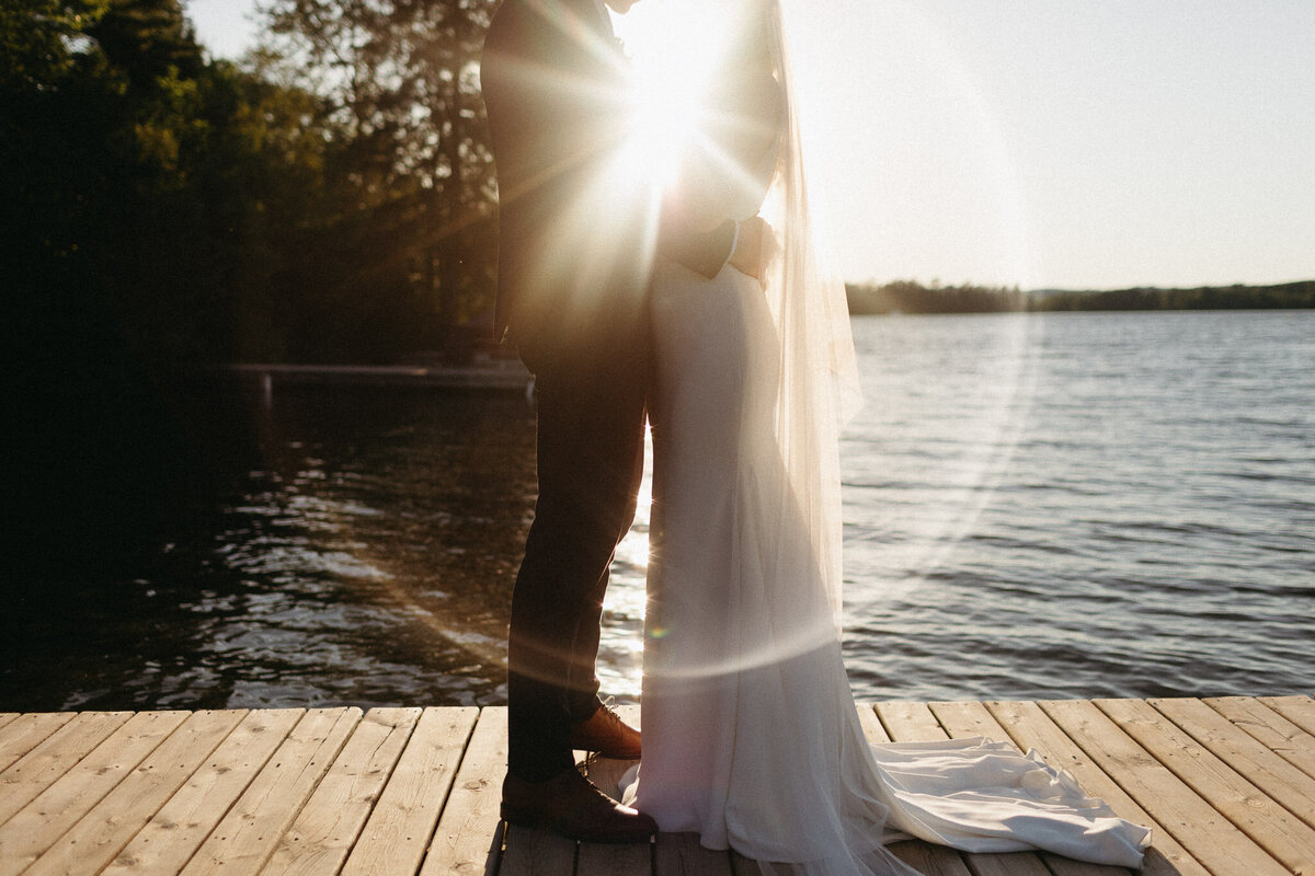 A couple stands embracing on a wooden dock by a lake, with the sun shining brightly between them, creating a lens flare effect. It's an Muskoka wedding photographer's dream moment, capturing love and nature's brilliance in perfect harmony.