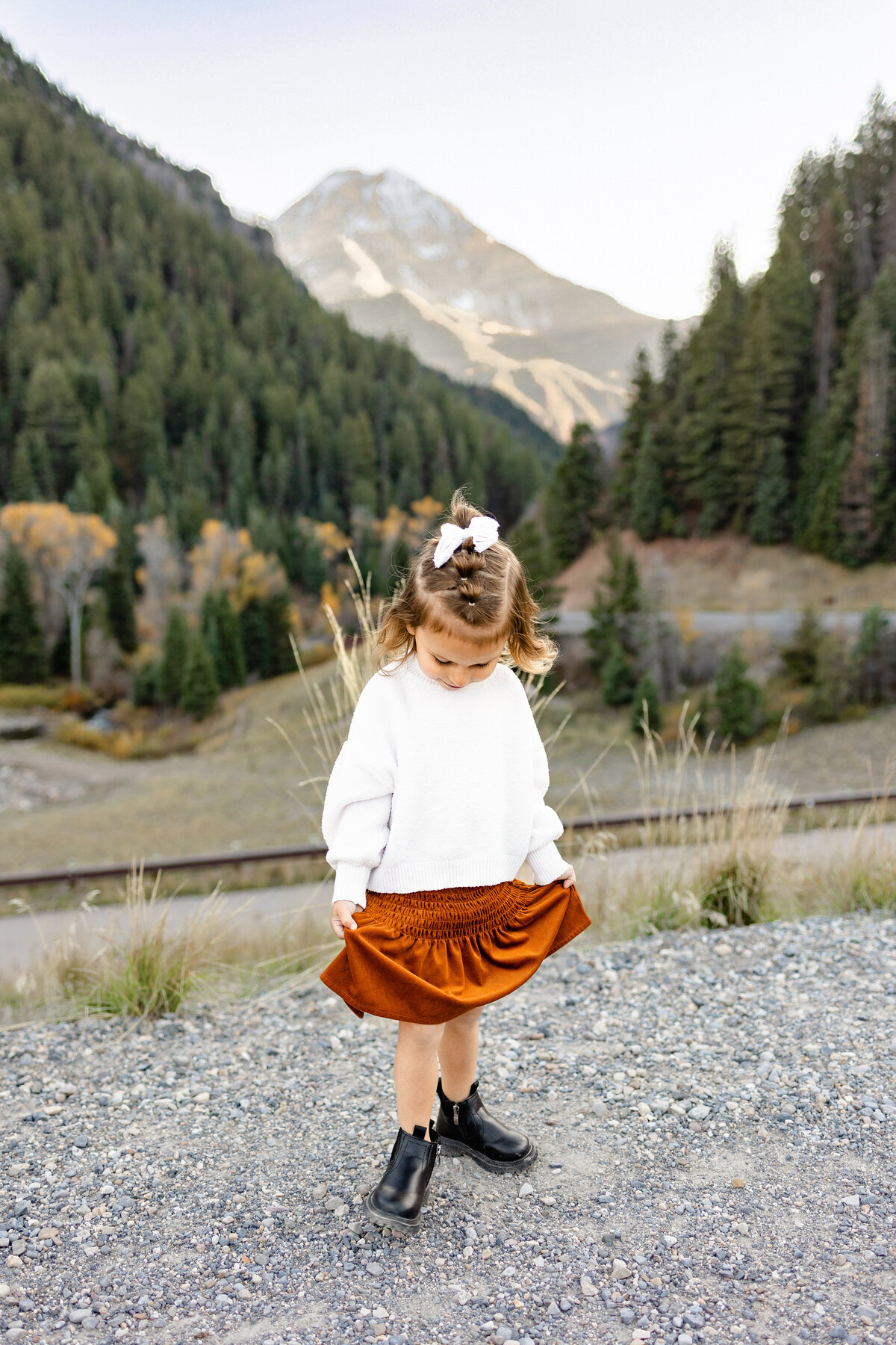 A young girl in a white sweater and rust skirt playfully twirls while standing on a rocky path, with towering evergreens and a snow-capped mountain in the background.