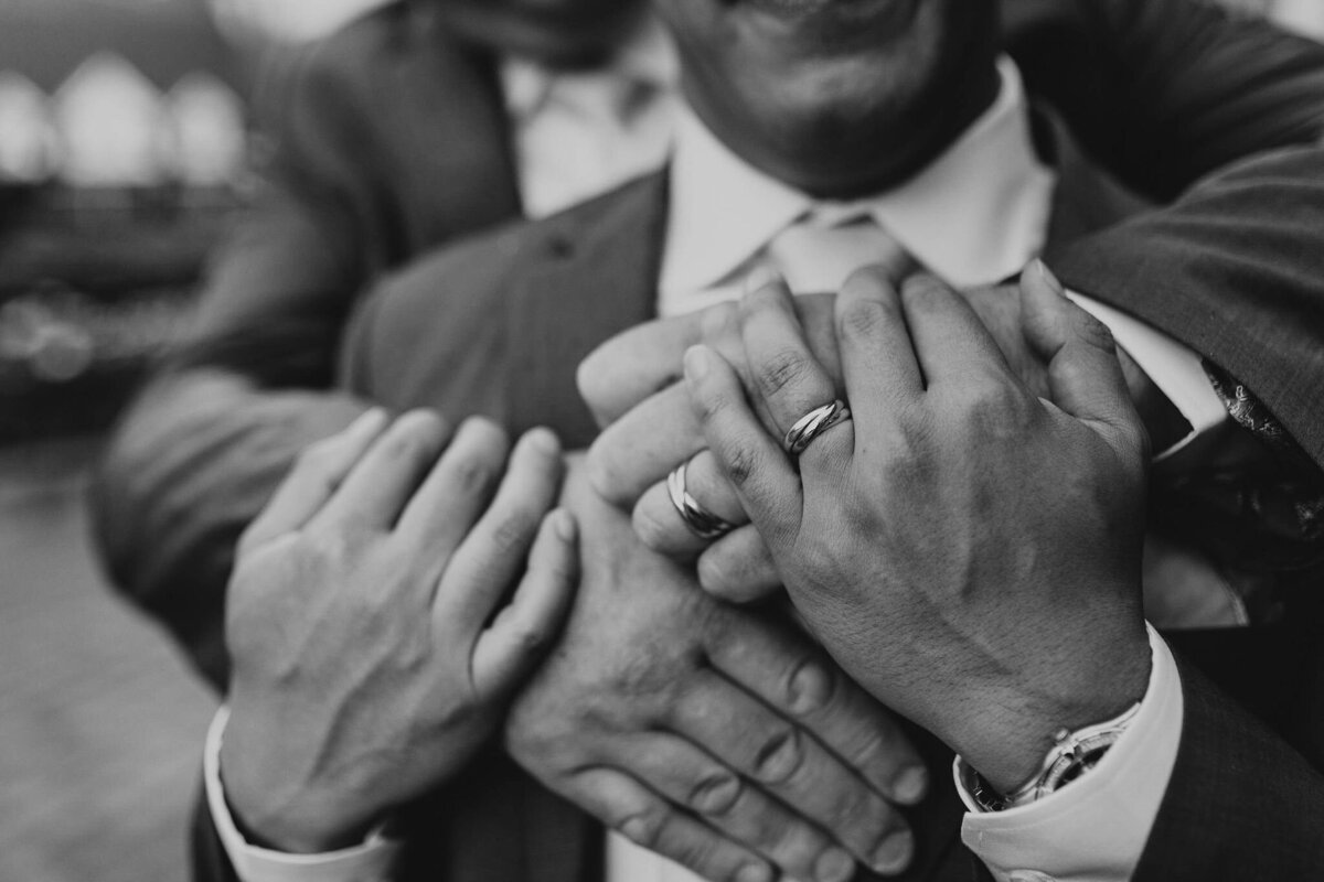 Black and white photo of three people wearing suits, each placing their hands on each other's arms, showcasing rings on their fingers—a modern-day fairytale wedding captured at the Banff Springs Hotel.