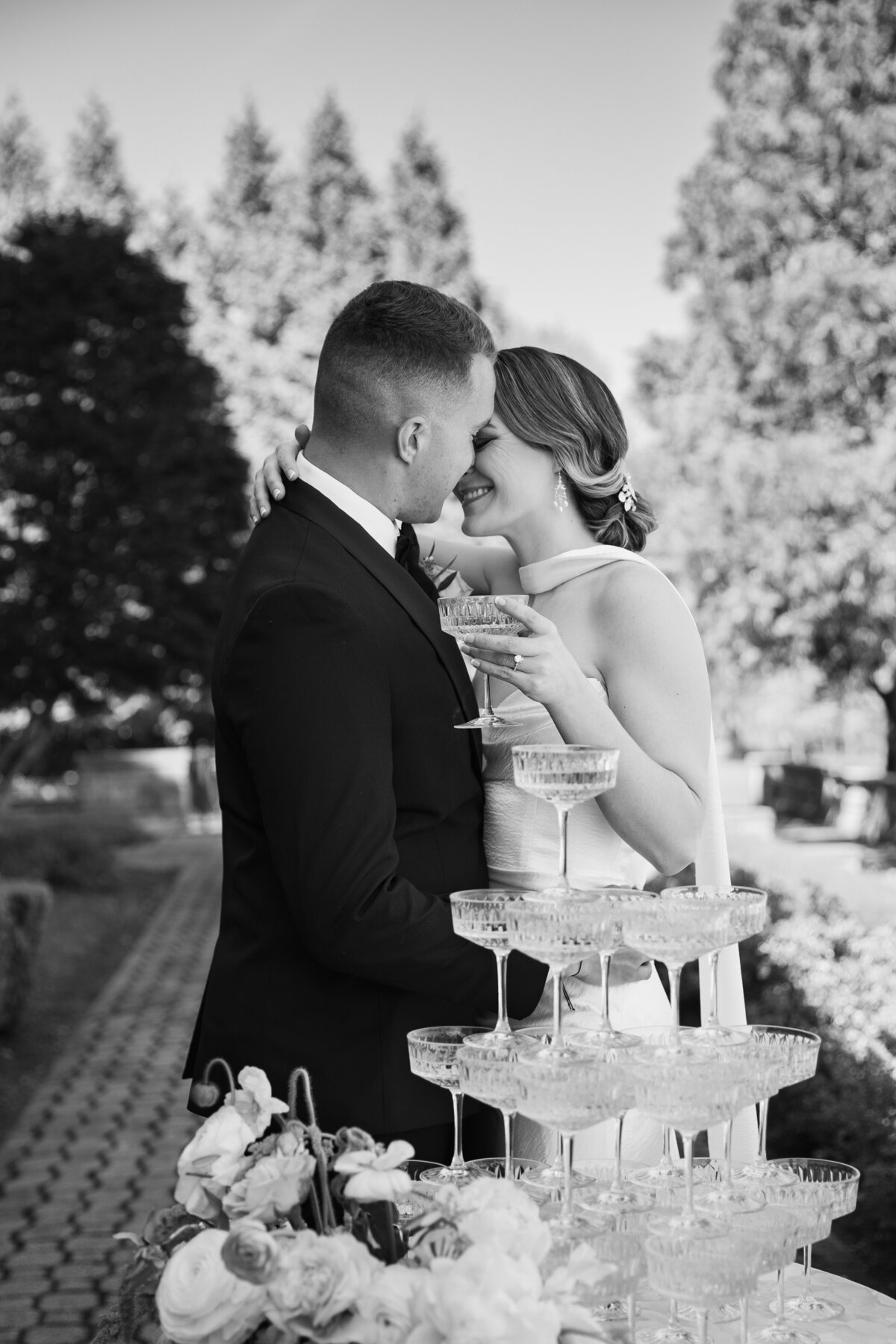 Aldrich Mansion Wedding Photos | Kelsey Sheehan Photography Timeless Rhode Island Weddings | A couple in wedding attire shares an intimate moment, smiling and holding champagne glasses by a tower of glasses. Sunlit garden setting suggests joy and celebration.