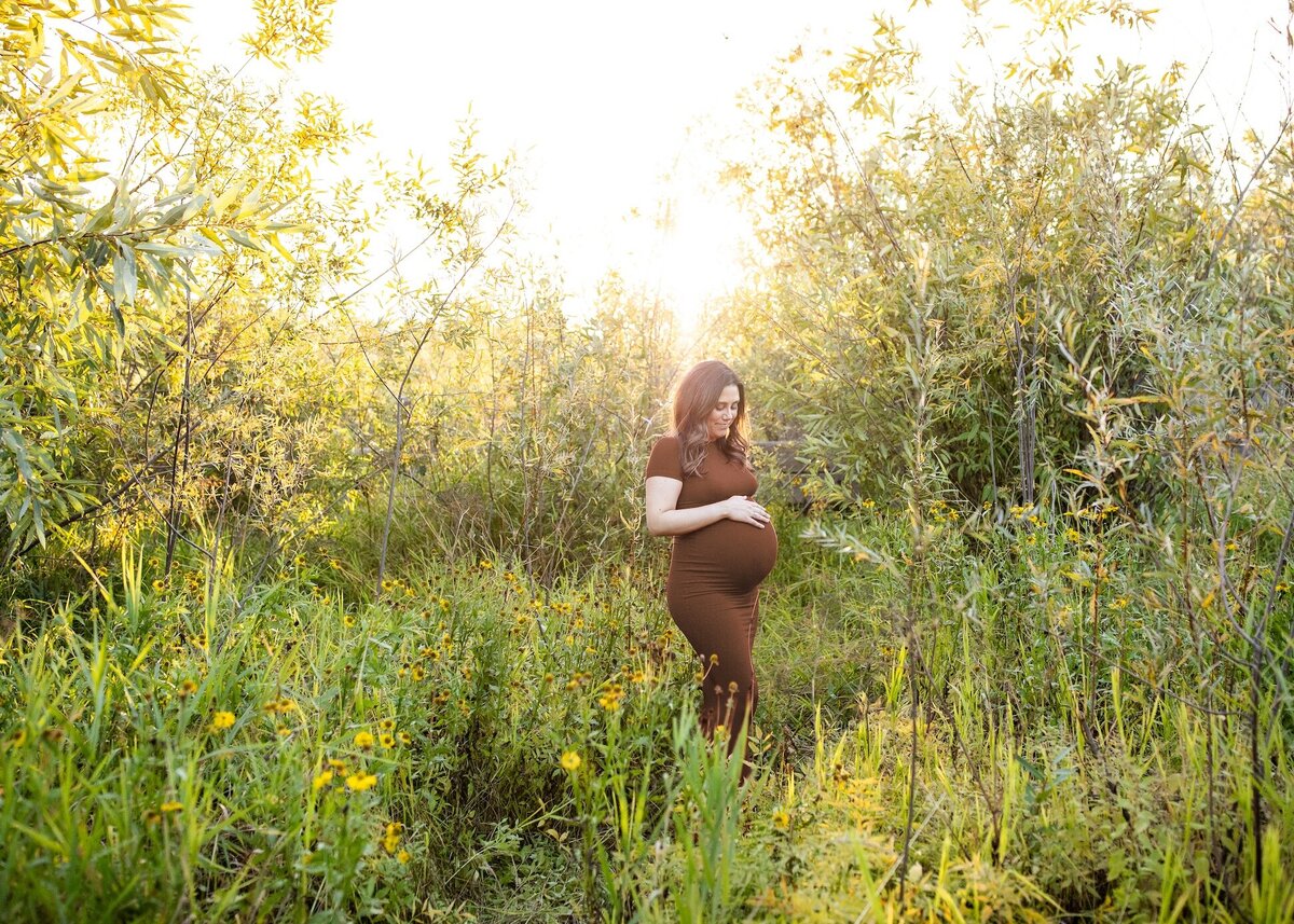 Mom standing in tall grass at sunset in brown maternity dress.