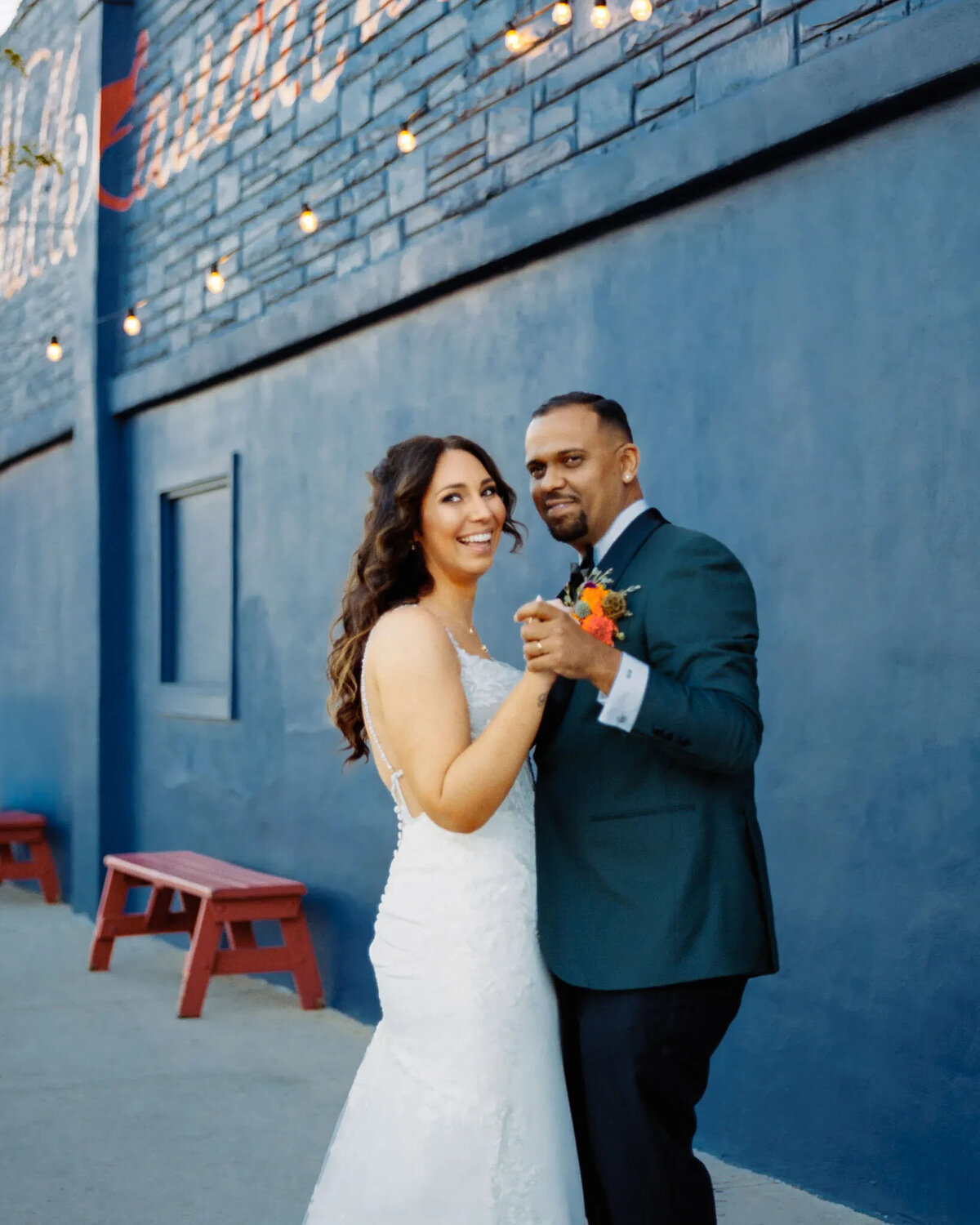 A bride in a white dress and groom in a dark suit smile while dancing outside by a navy blue wall with string lights and red benches, beautifully captured by an NJ wedding photographer.