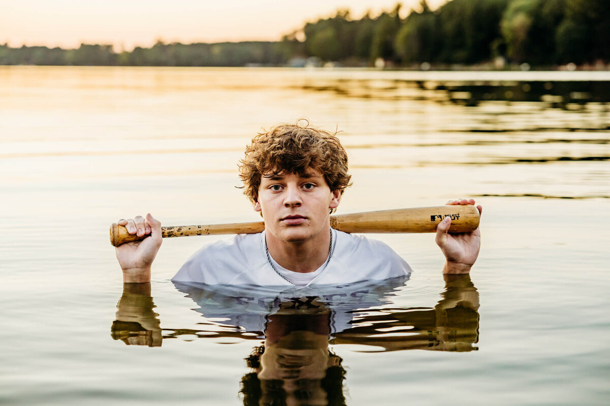 brown haired high school boy standing in the water up to his shoulders as he holds a bat on his shoulders in a lake near Green Bay.