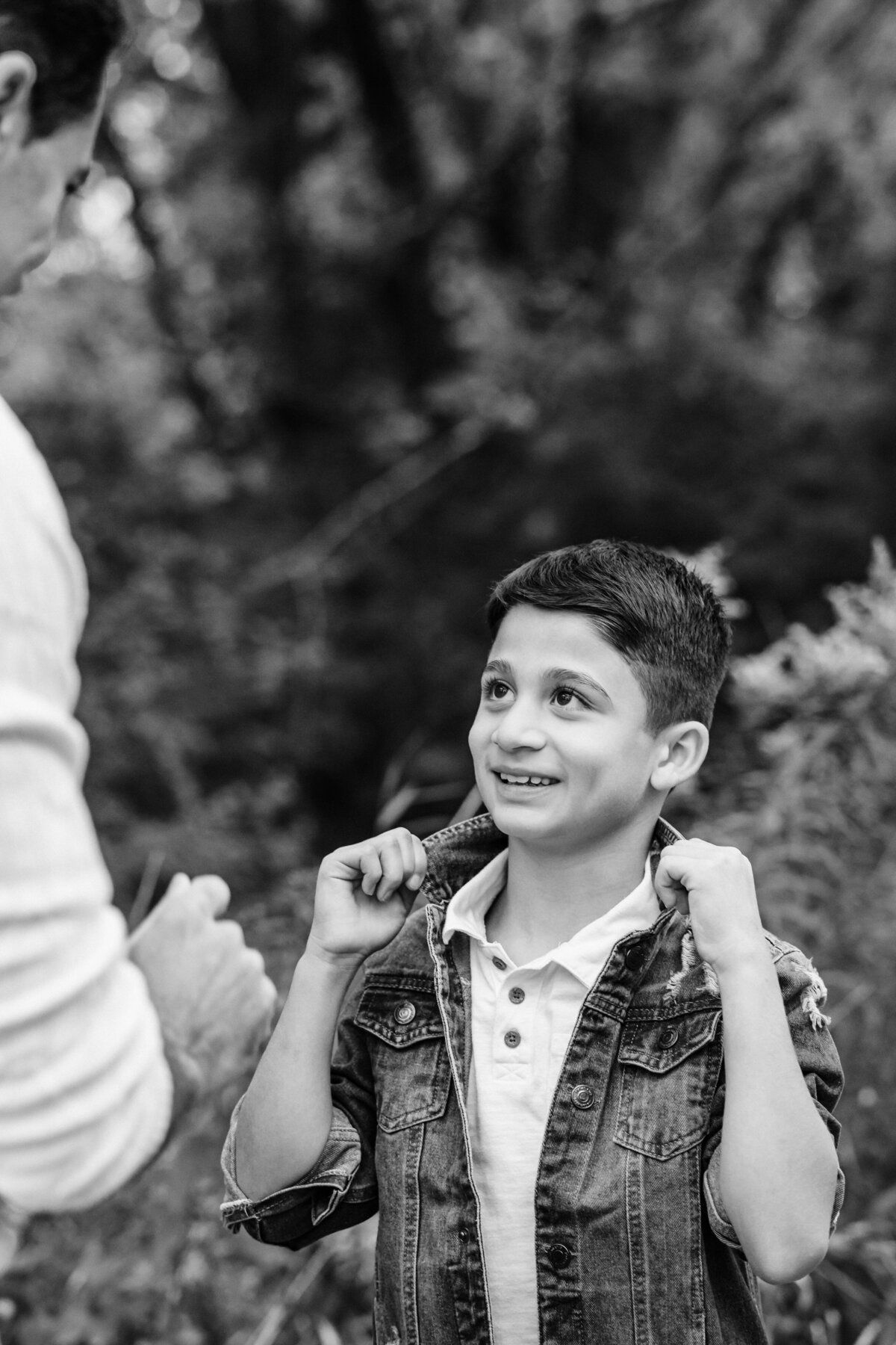 A black-and-white photo of a young boy smiling up at an adult, likely his father, while adjusting his denim jacket. The boy looks happy and engaged, with a background of trees and foliage.