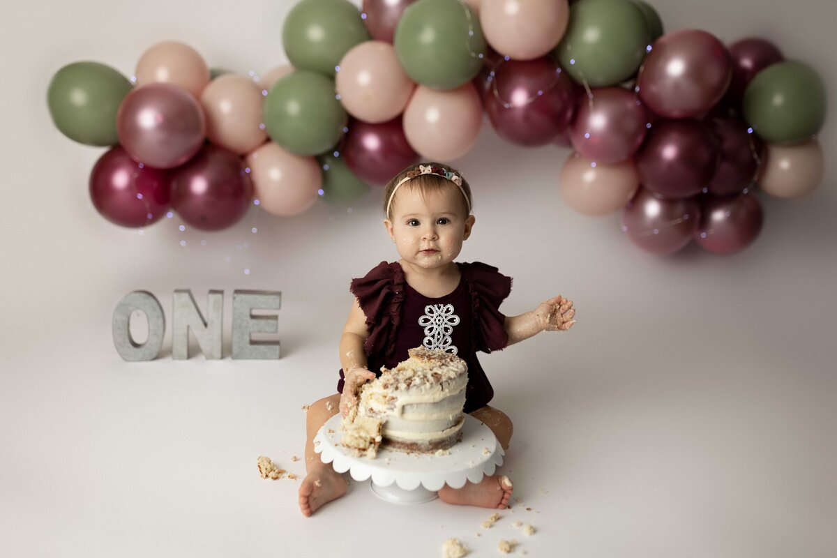 Studio Cake Smash - Baby in black outfit with messy cake in front of them and purple and green balloon arch behind them.