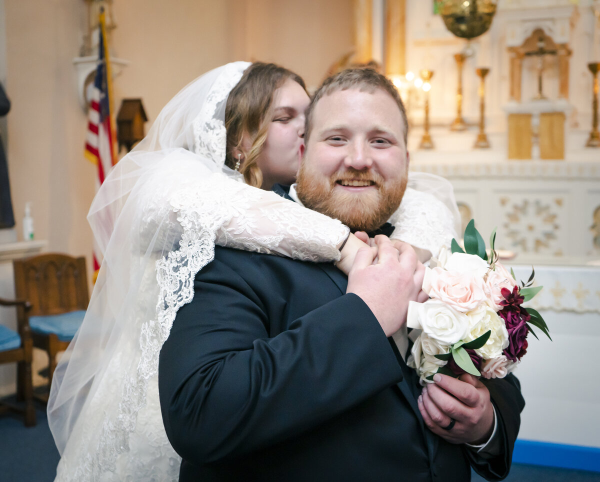 Photo of a bride kissing her husband as he smiles at the camera