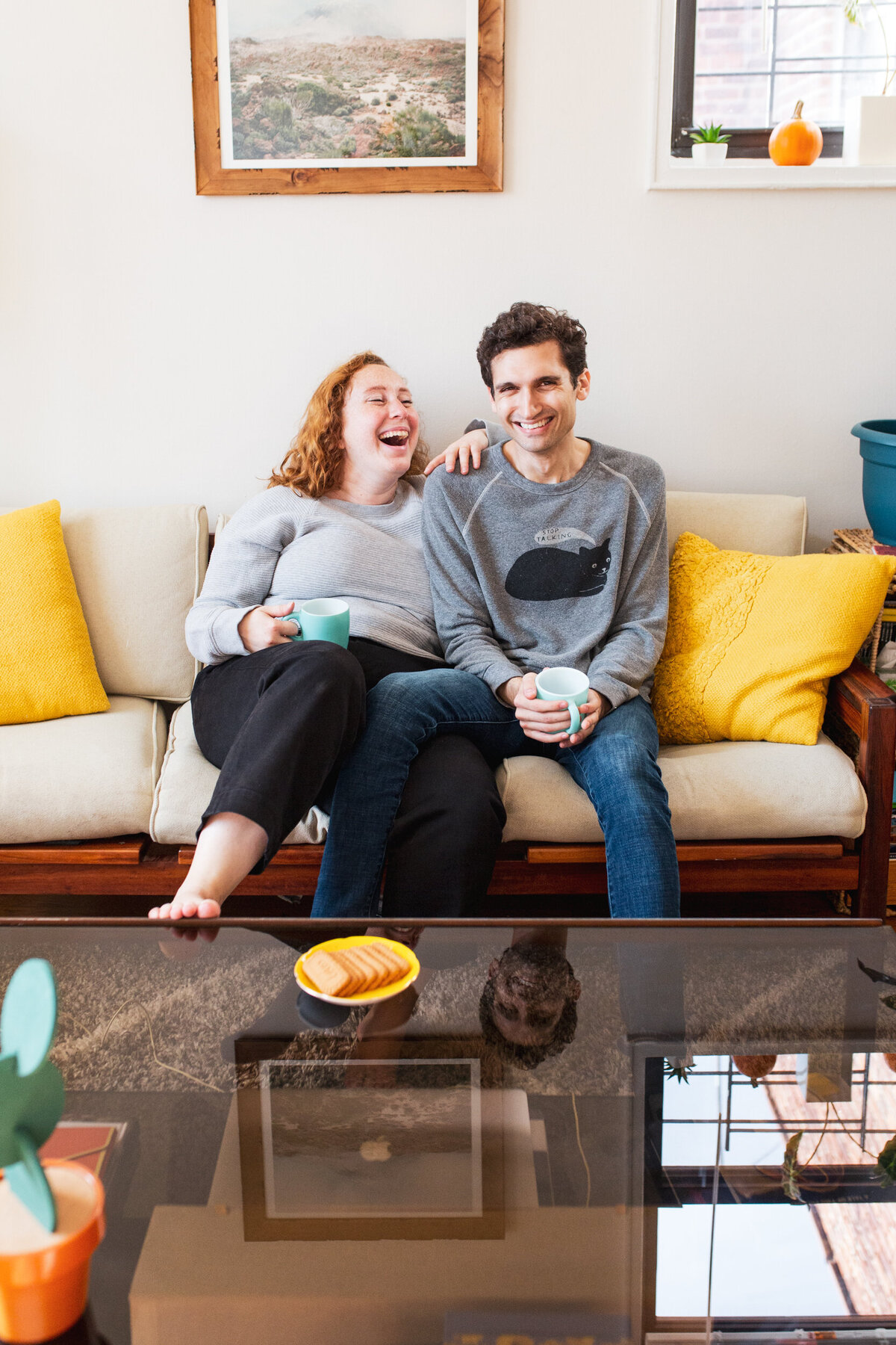 Relaxed, candid in-home couples session in Queens, New York. A curvy woman sits on the couch next to her fiance and they laugh while drinking tea.