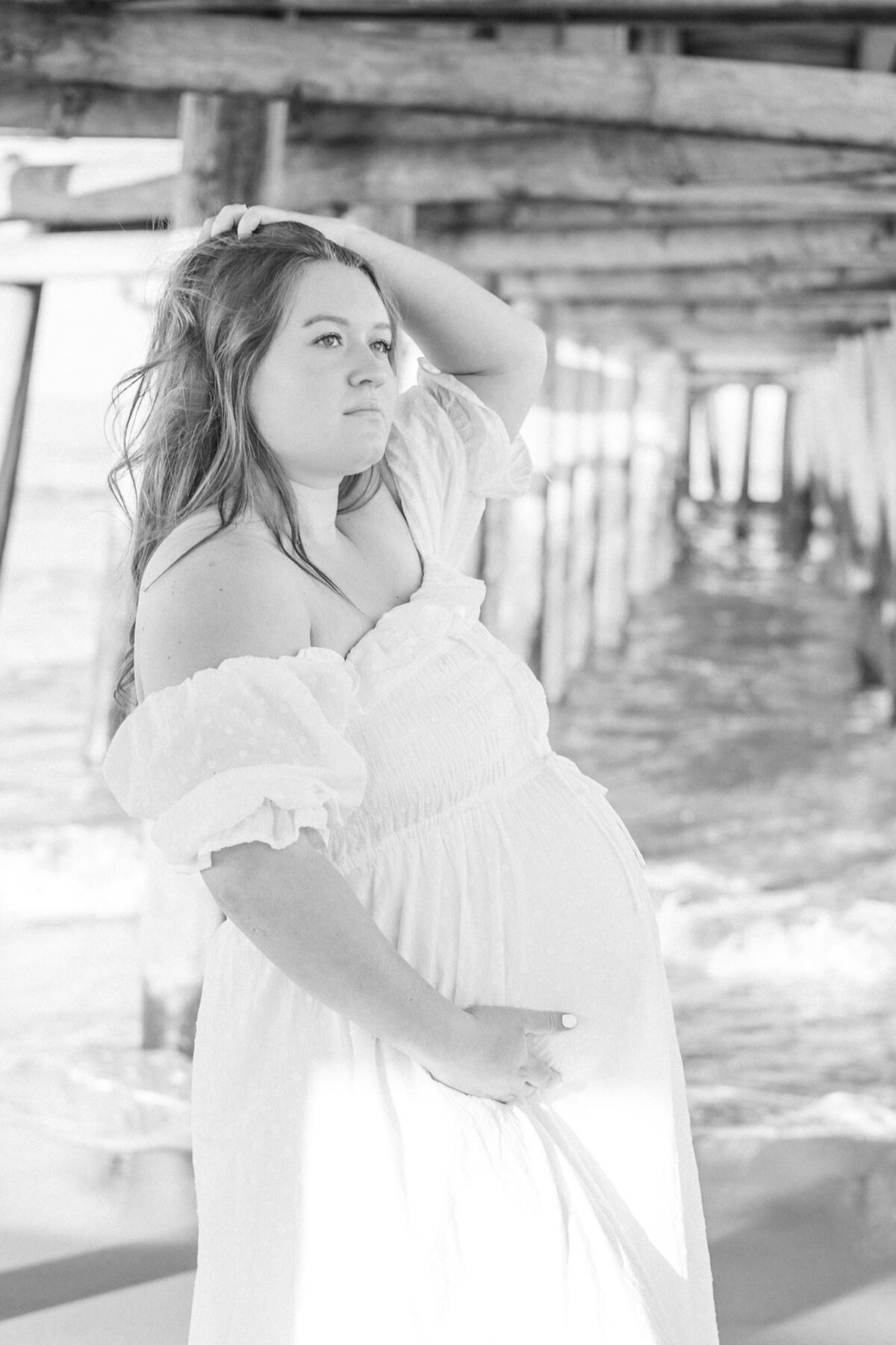 Black and White Portrait of a pregnant woman wearing a white off the shoulder dress, standing under the pier cradling her belly by Virginia Beach Maternity Photographer Mary Eleanor Photography