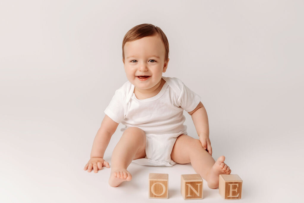 Baby boy in white onesie celebrates milestone first birthday with wooden alphabet blocks spelling ONE on clean white backdrop