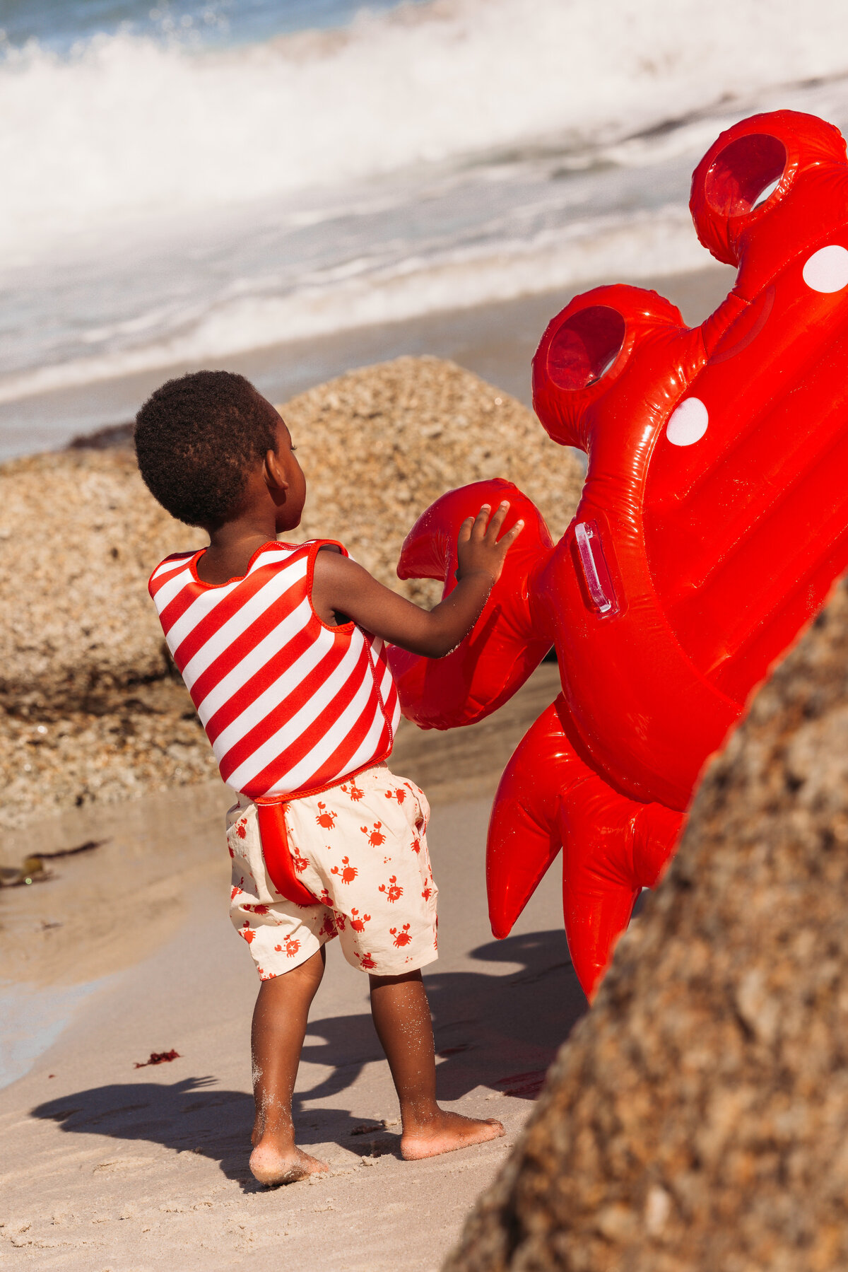 Young boy in a red-and-white striped swim vest and crab-print swim shorts playing with a large inflatable red crab on the beach in Cape Town.