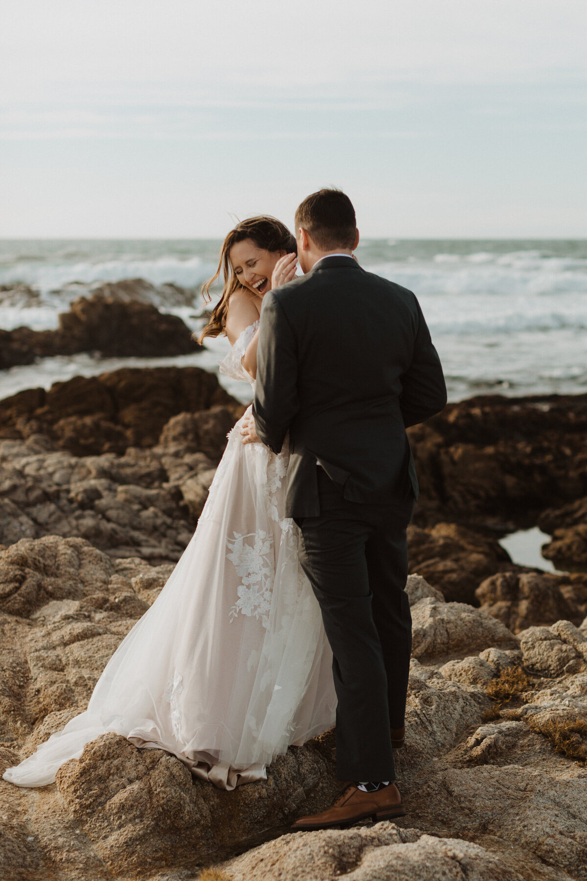 Bride laughing and facing groom while standing on rocky ocean coast