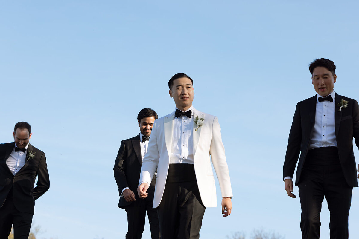 Groom in white against blue afternoon sky walks near his groomsmen at Trinity View Farm in Franklin, TN.