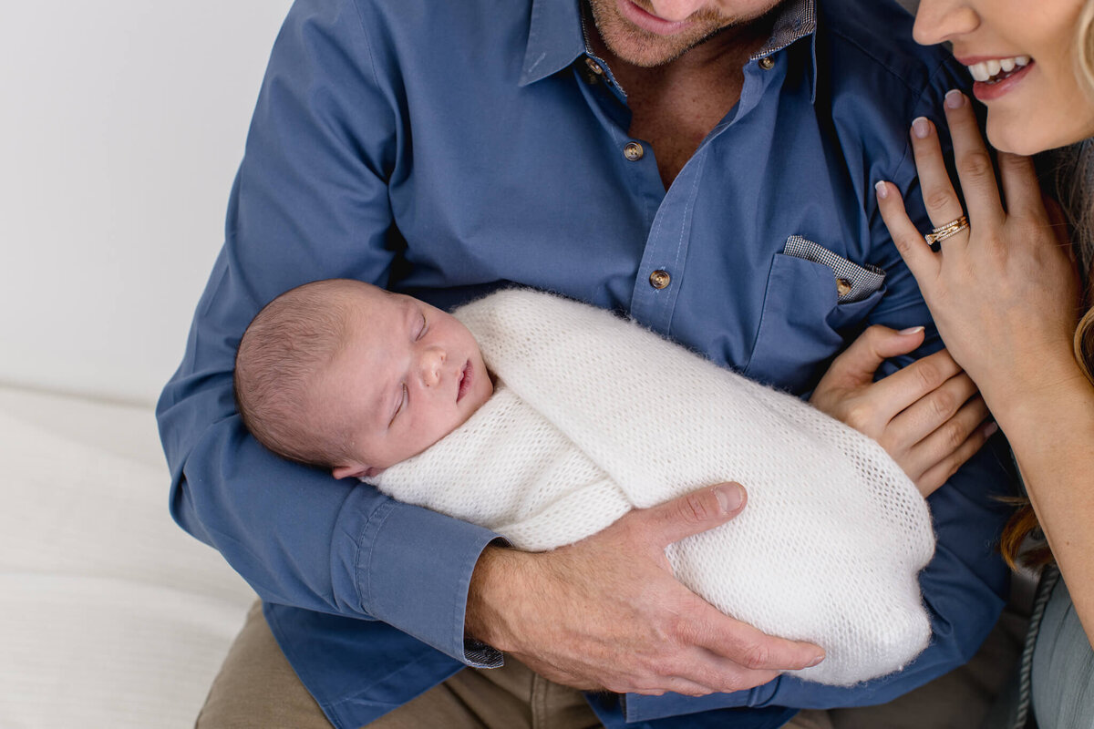 A baby is wrapped in a white blanket, sleeping peacefully in the arms of a man wearing a blue shirt. A woman gently touches the mans arm, smiling.