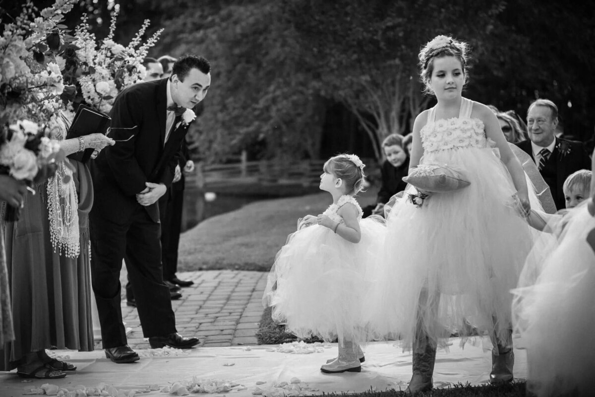 A groom leaning down and talking to a flower girl as she walks down the aisle.