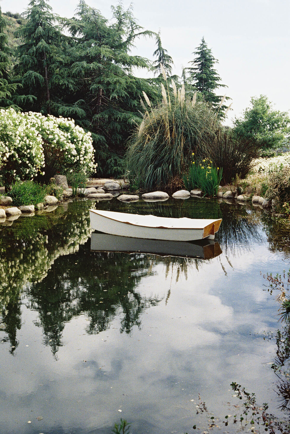 A small white boat floats on a serene pond surrounded by lush greenery and rocks, with trees and the sky reflected in the water, creating a peaceful atmosphere at Serendipity Garden in Oak Glen.