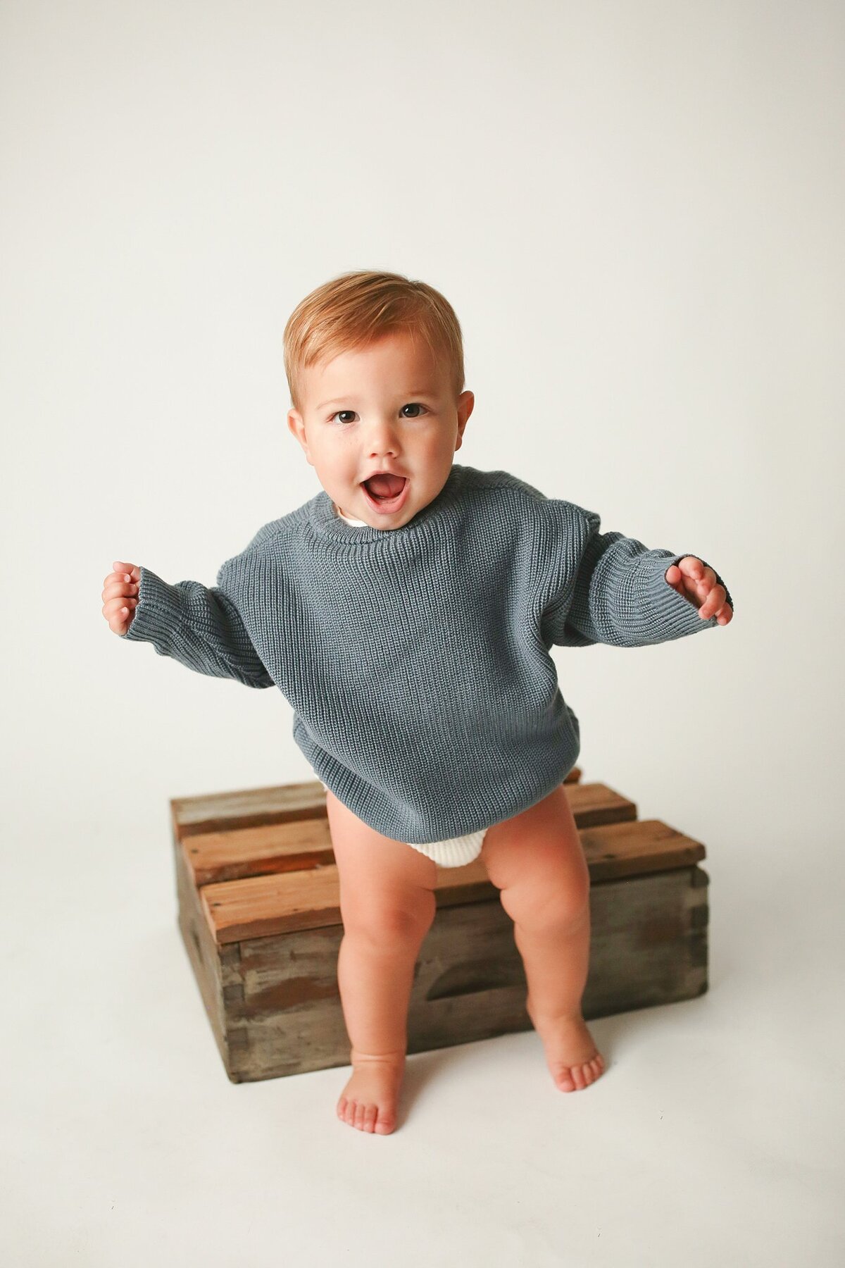 A-smiling-baby-wearing-a-cozy-blue-sweater-standing-on-a-wooden-crate-during-a-milestone-studio-photo-session-at-Studio-1432-on-Main-Street-in-Speedway-Indiana-near-the-Indianapolis-Motor-Speedway-perfect-for-documenting-baby’s-growth-and-milestones