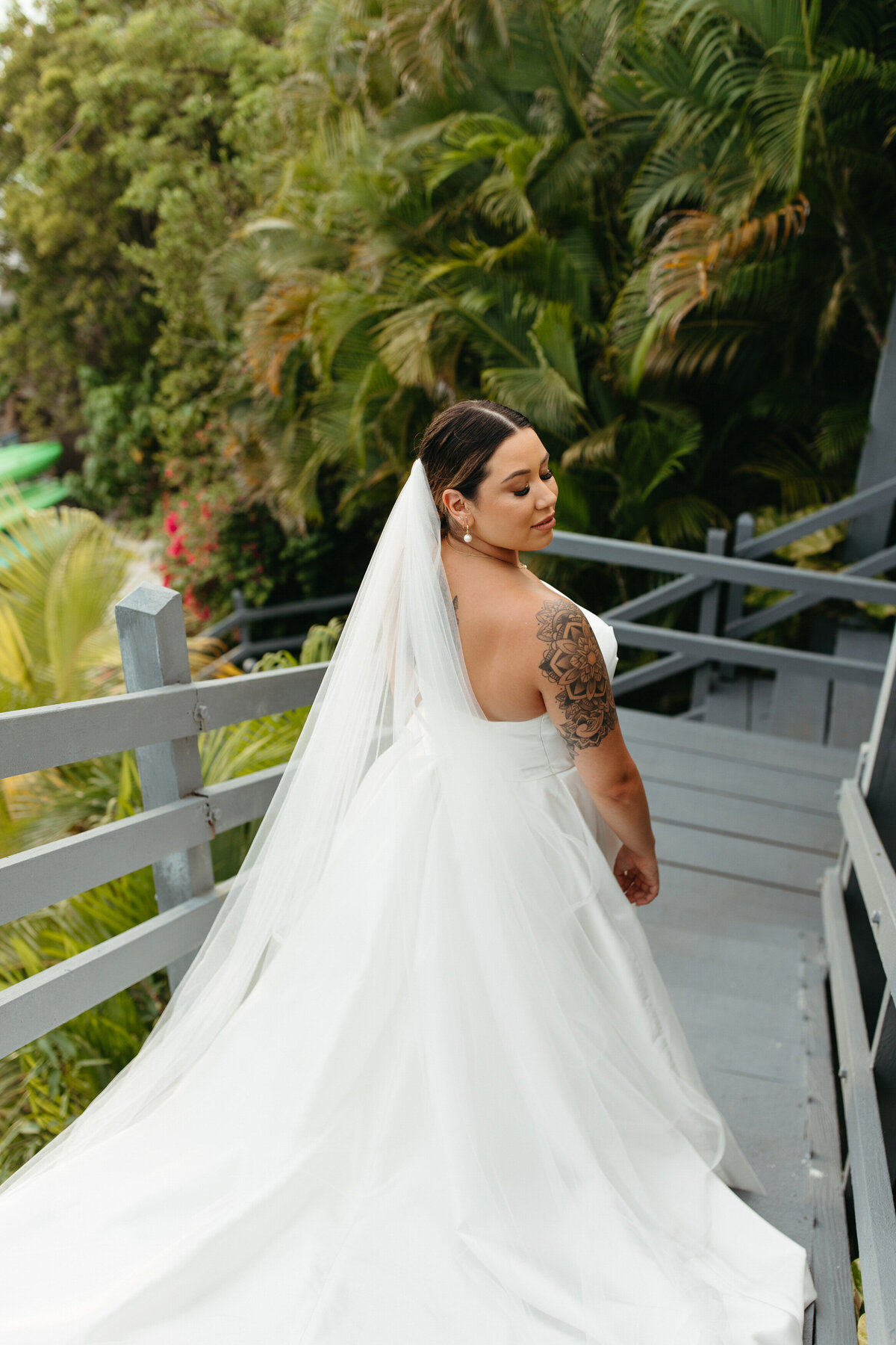 Bride in a flowing white dress gazing back on a wooden walkway, surrounded by rich foliage
