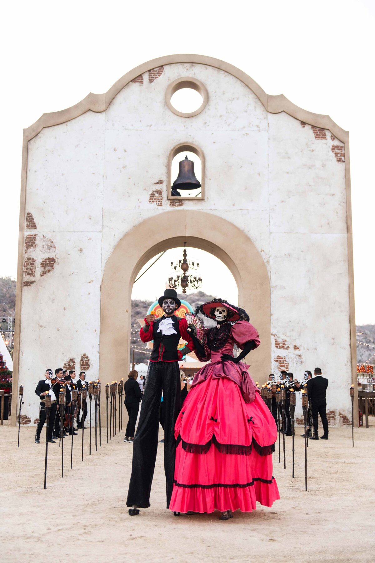 Day of the Dead characters and mariachis pose in front of bell tower set wall to welcome guests