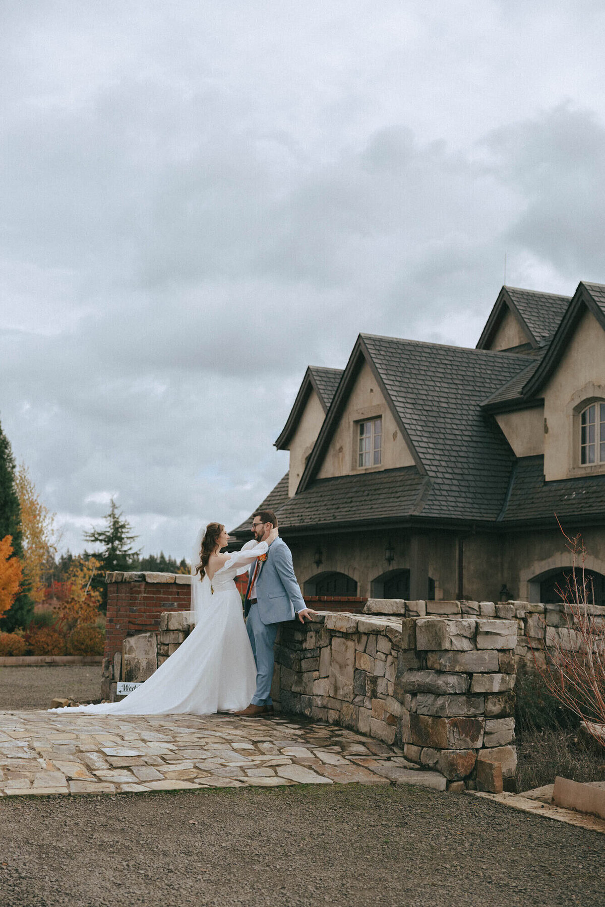 A bride and groom embrace while the groom leans against a rock wall at the Chateau wedding venue in Oregon.