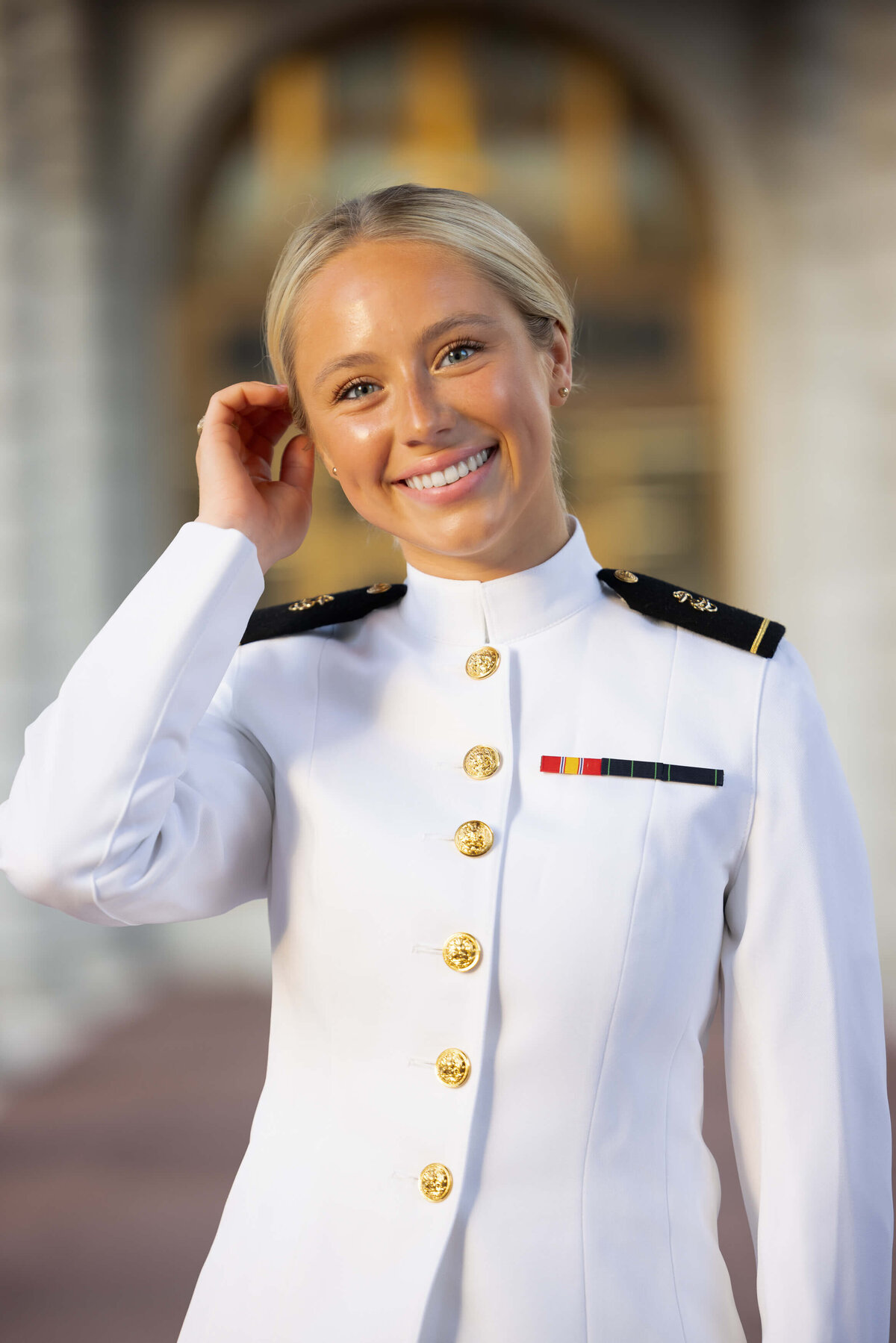 USNA Midshipman tucks blonde hair behind her ear and smiles in front of golden doors