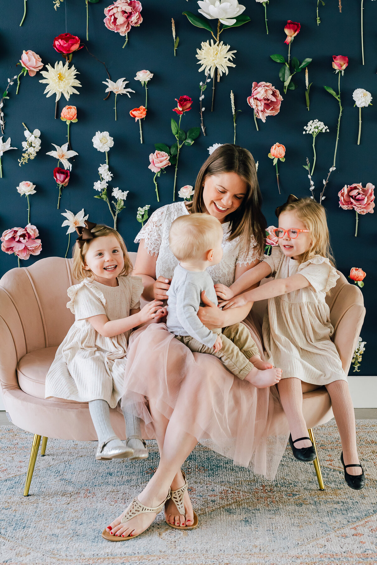 denver family photos with colorado wedding photographers sitting on a pink bench with her three children smiling and cuddling with one another in front of a floral backdrop