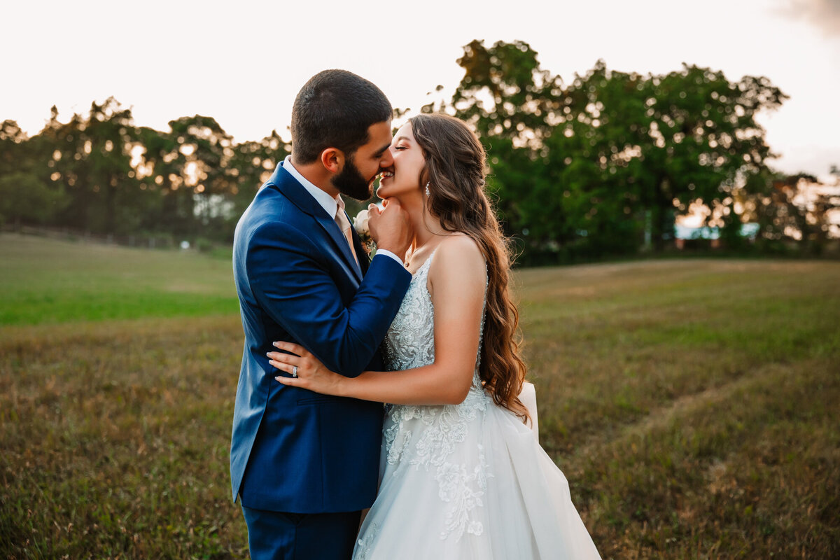 grooms takes brides chin to kiss her in a field at sunset