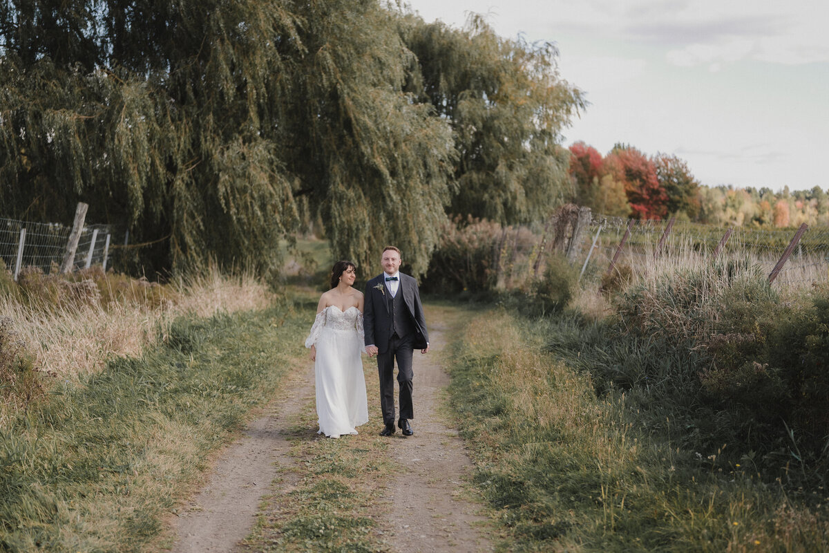 Couple de mariés marchant main dans la main dans un sentier champêtre au Vignoble de la Bauge, en pleine nature automnale, capturé par Jeff Gagnon photographe.