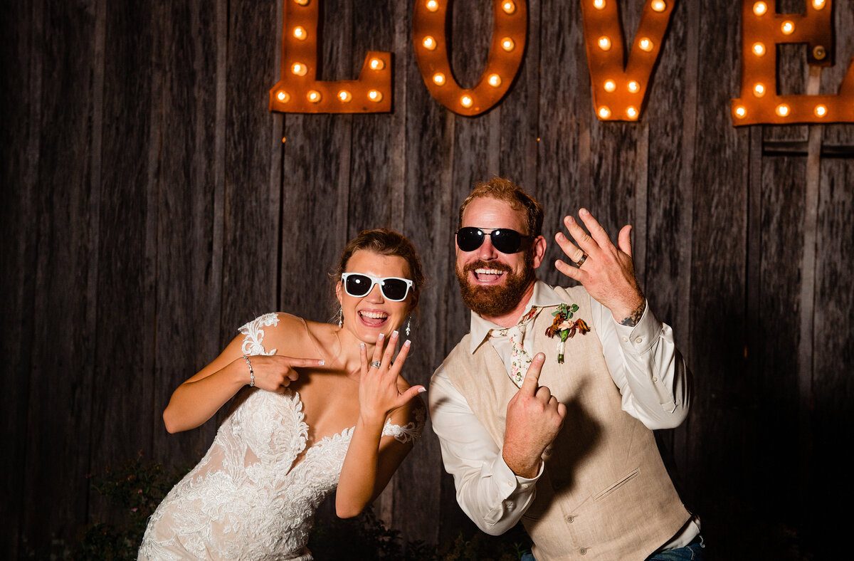 Newlyweds holding up hands showing off their wedding hands and wearing sunglasses at night in front of the Love letter sign at Drakewood Farm