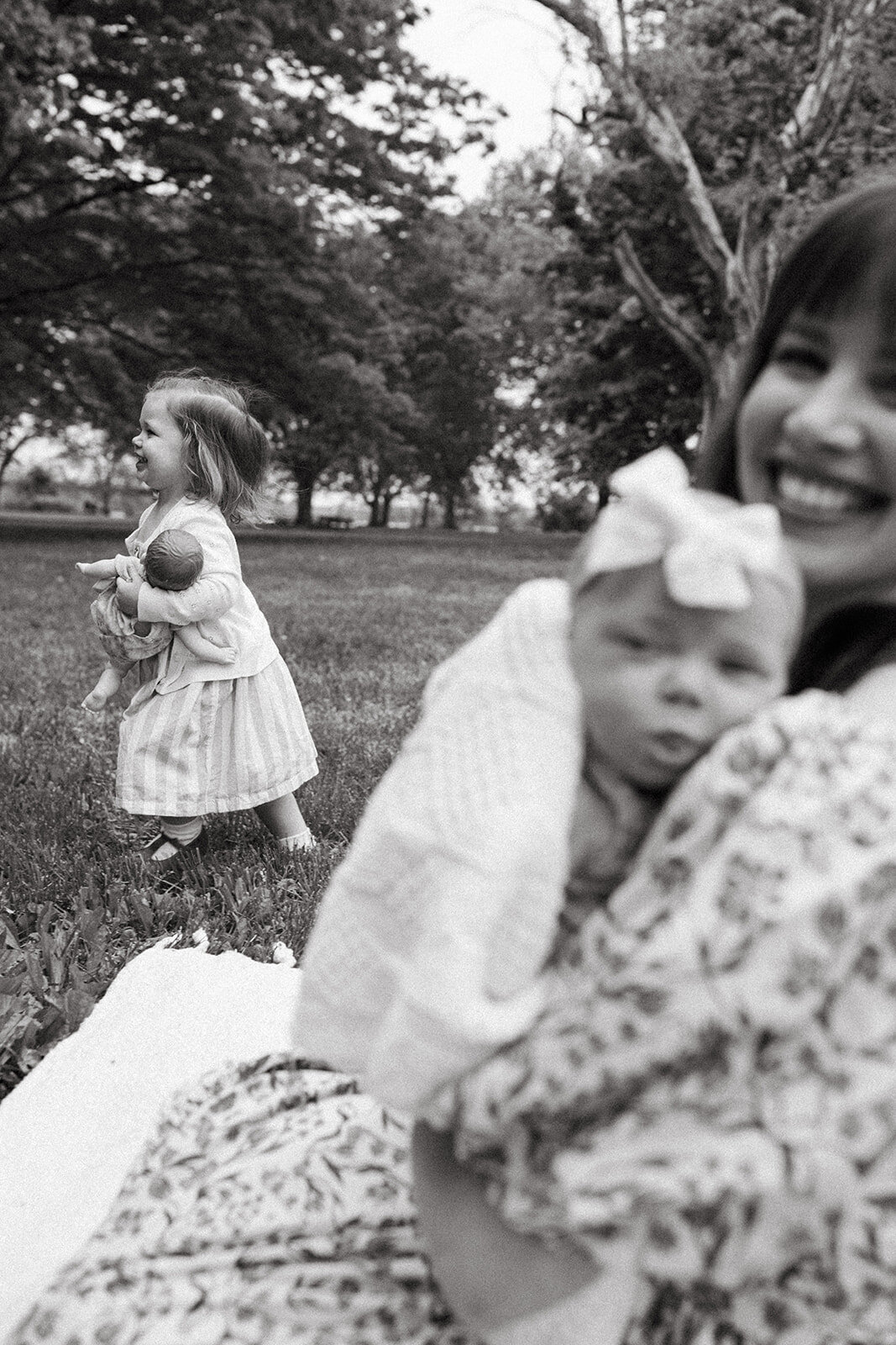 Little girl running in a yard with a baby doll in her arms while her mother watches from the foreground

